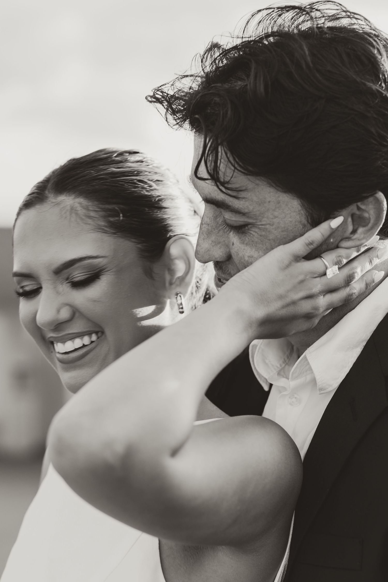 Black and white photo of a smiling bride holding groom's face tenderly during a romantic wedding moment.