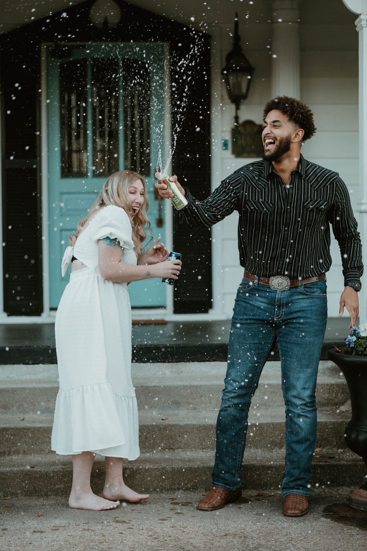 Couple celebrating with champagne spray outside a home, woman in white dress and man in striped shirt and jeans.