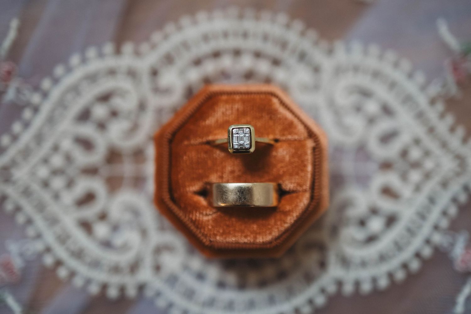 A diamond engagement ring sits in an orange octagonal ring box on white lace fabric.