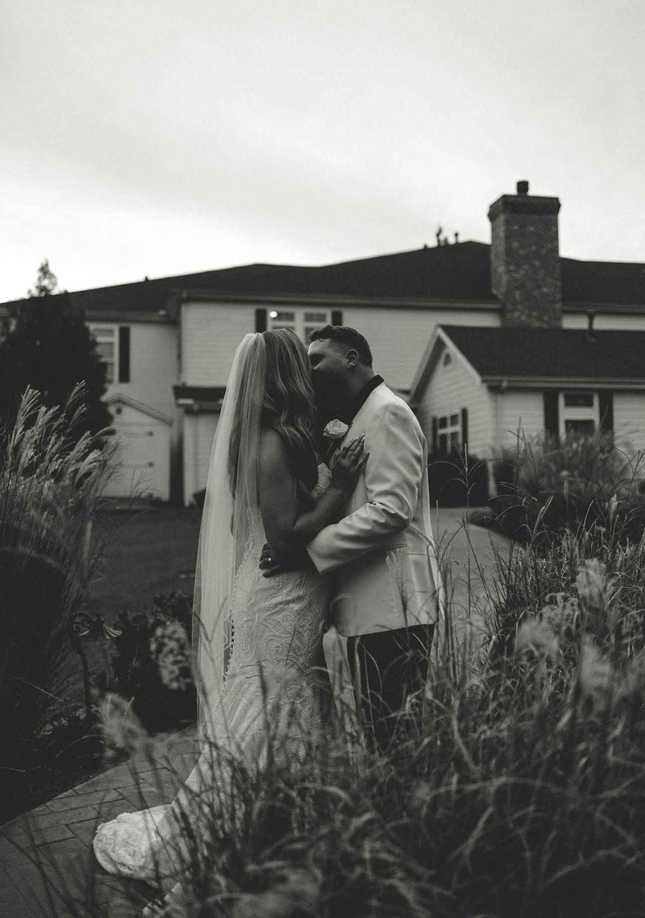 Black and white photo of a bride and groom sharing a kiss in a garden outside a white house on their wedding day.