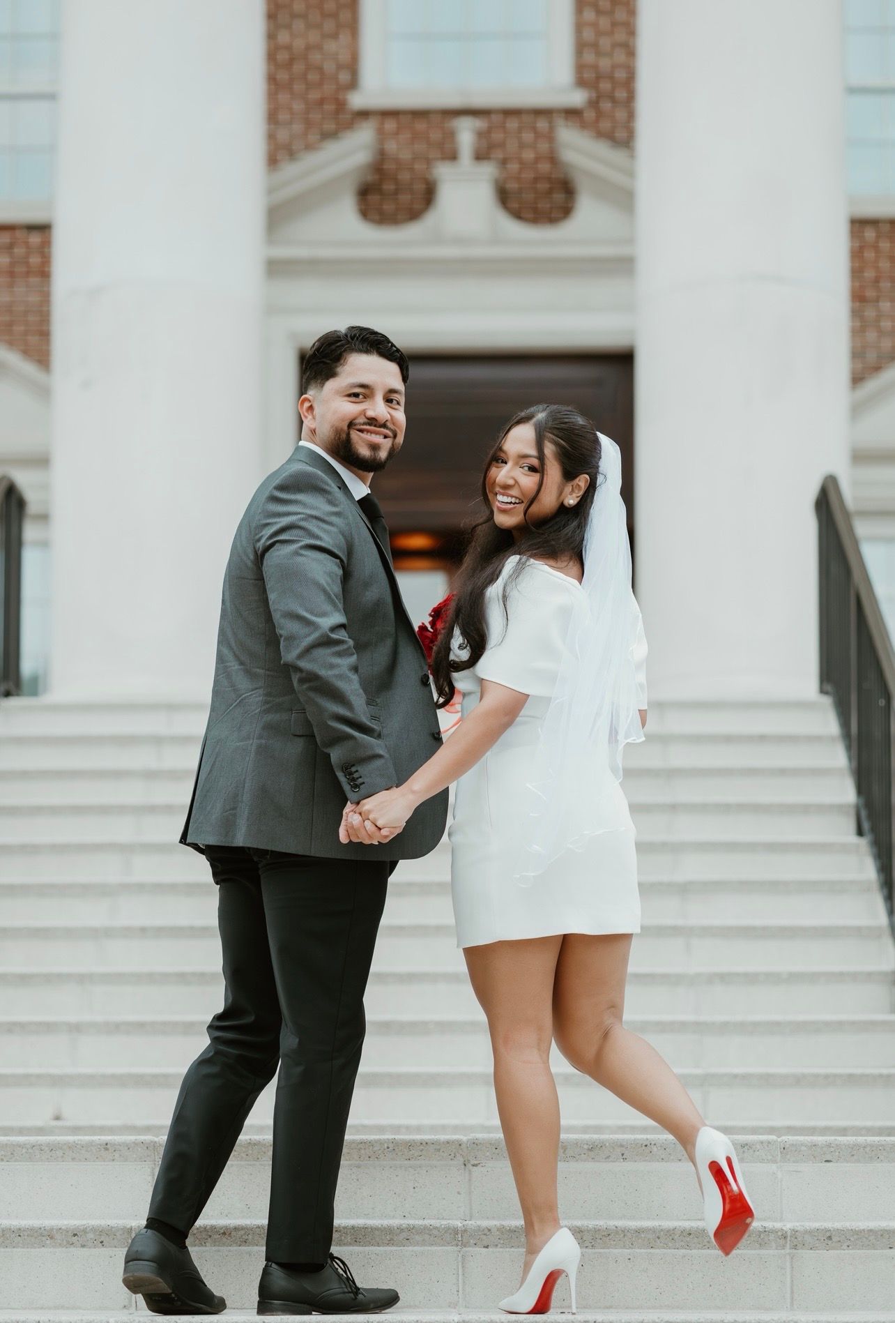 Smiling newlywed couple holding hands on church steps, bride in white mini dress with veil and red-sole heels, groom in gray suit.