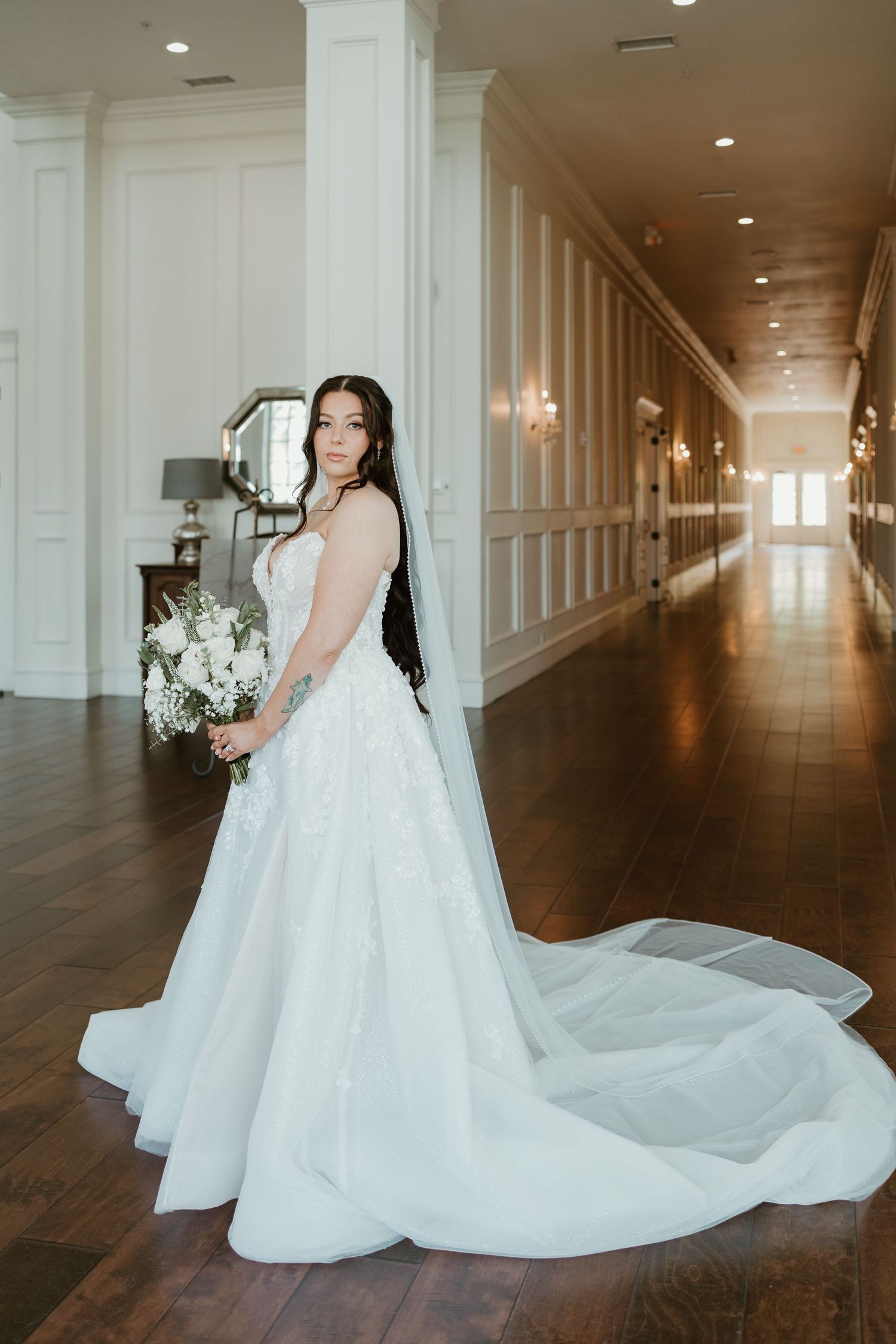 Bride in a white ballgown with lace details and long veil holds a bouquet in an elegant white hallway.