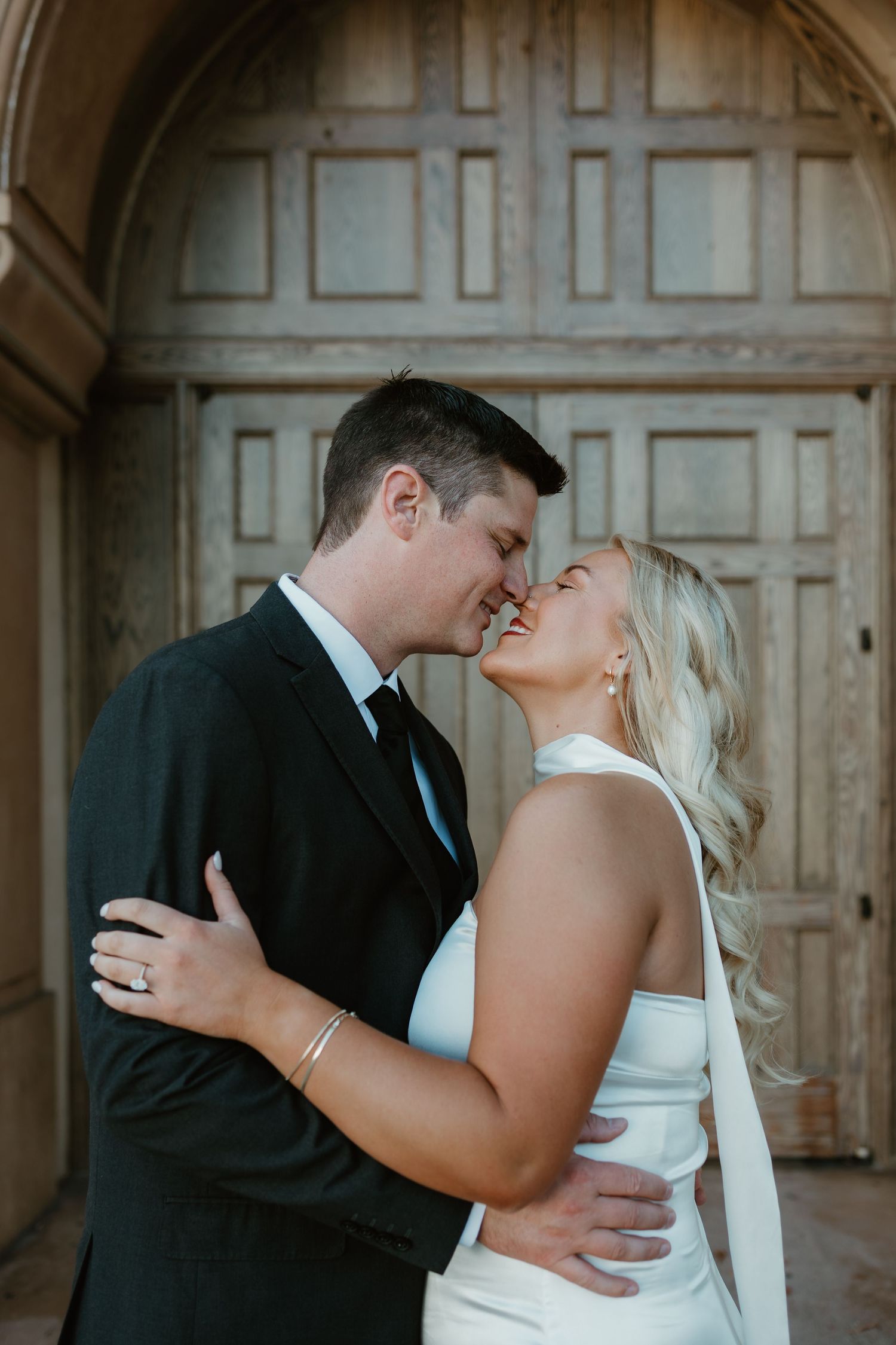 A bride and groom share a romantic almost-kiss moment in front of rustic wooden doors in wedding attire.
