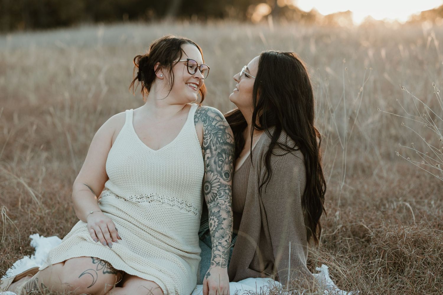 A romantic couple sits together in a sunlit meadow during golden hour with warm, hazy lighting.
