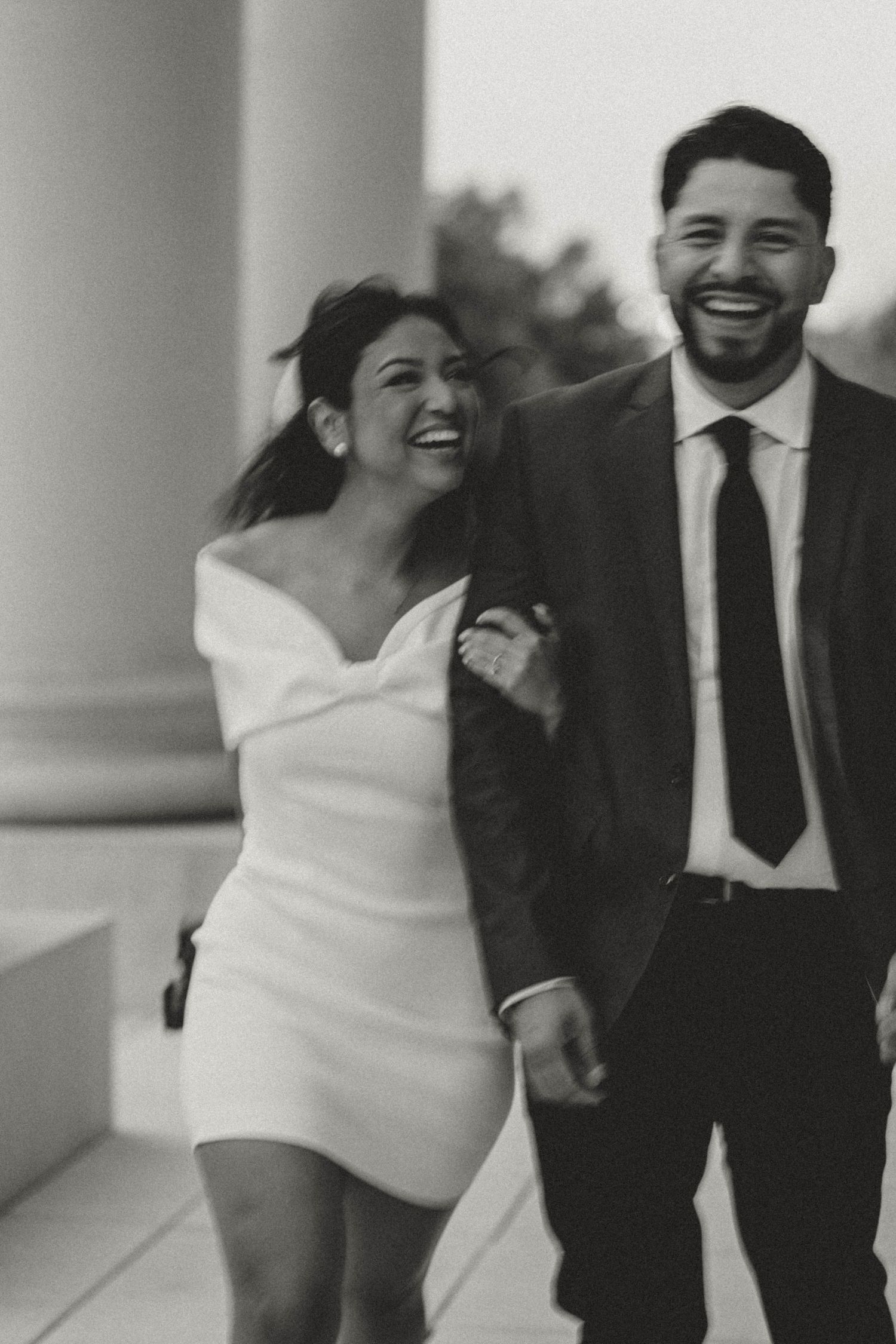 Black and white photo of a laughing couple in wedding attire walking together between columns.
