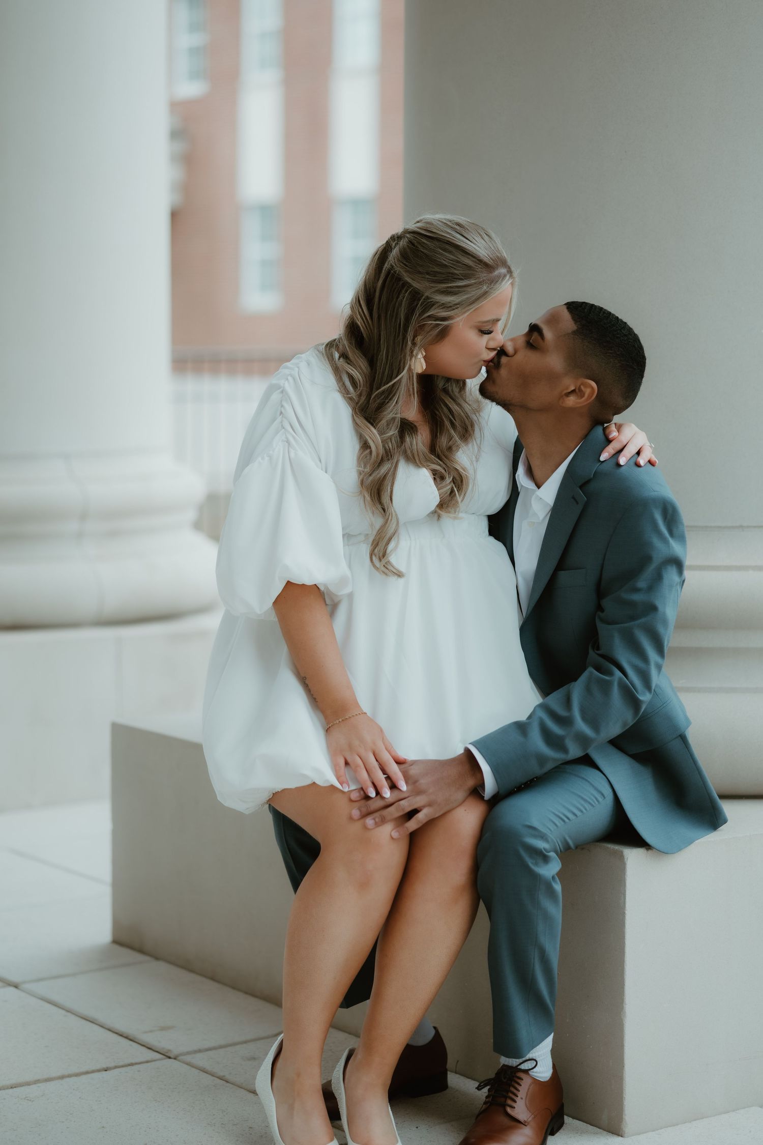 A romantic couple in formal wear share an intimate moment while sitting on a concrete ledge against white columns.