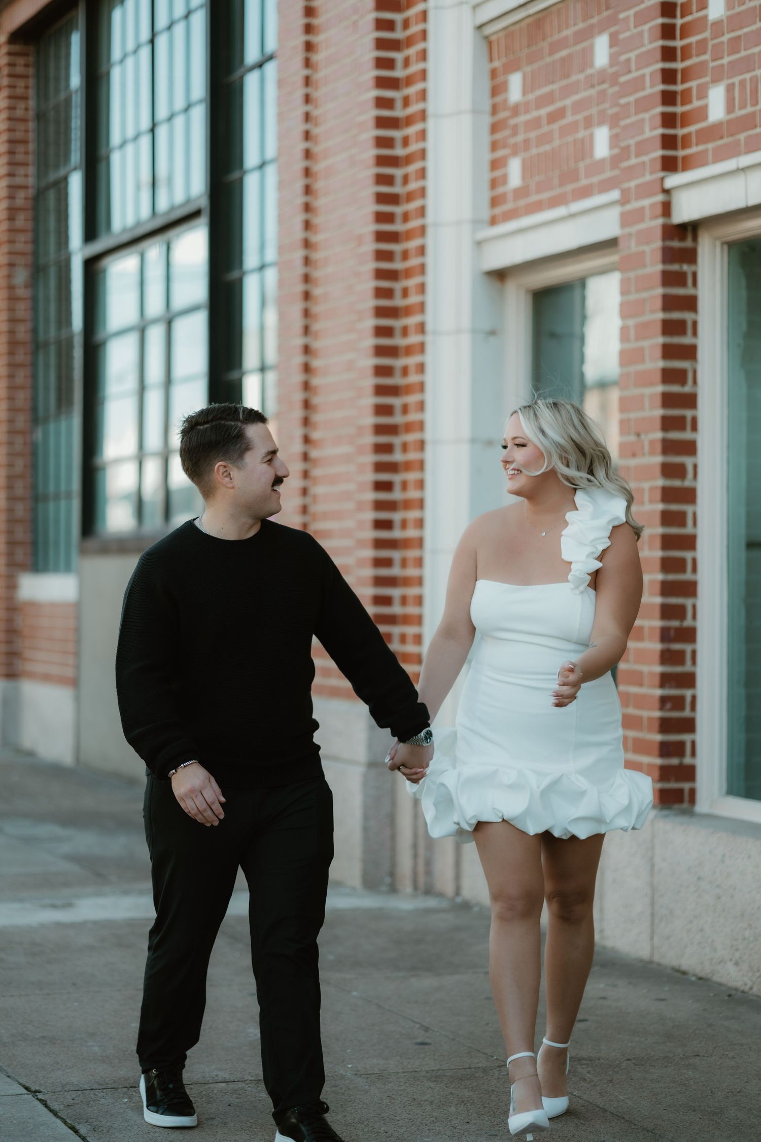 A couple in contrasting black and white outfits walk together holding hands along a brick building wall.