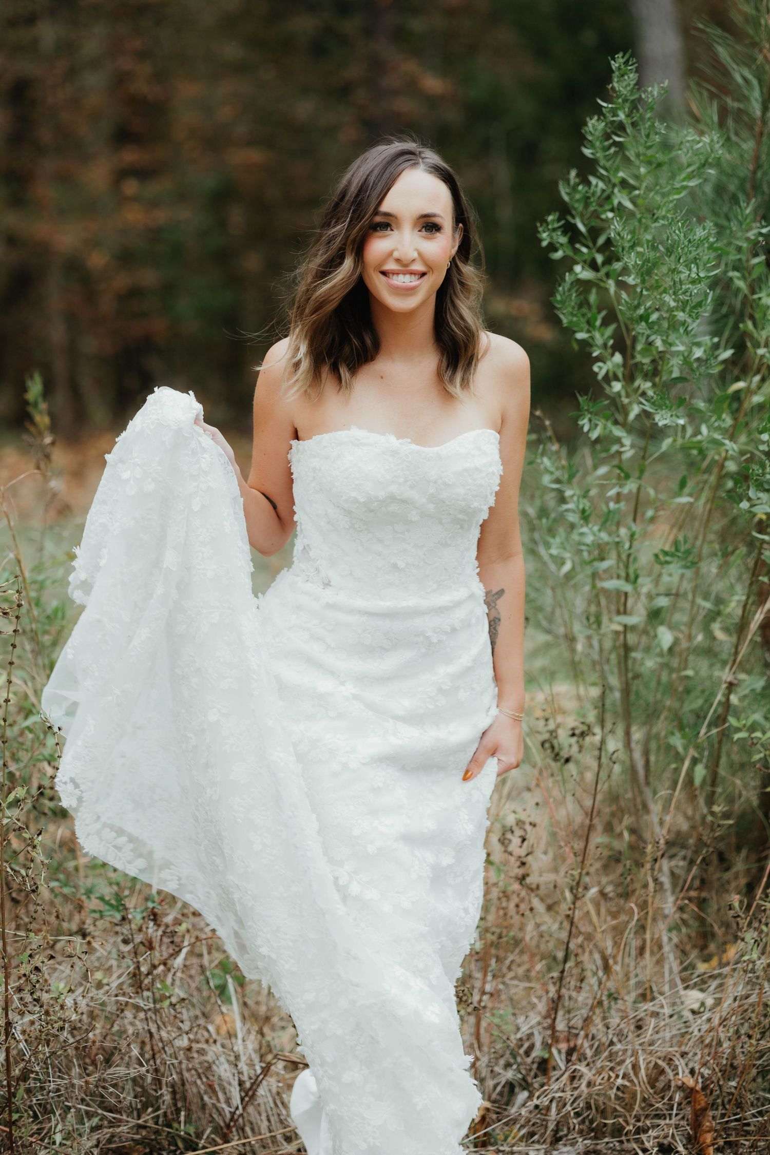 Smiling bride in strapless white lace wedding gown holding her flowing train outdoors in a natural field setting.