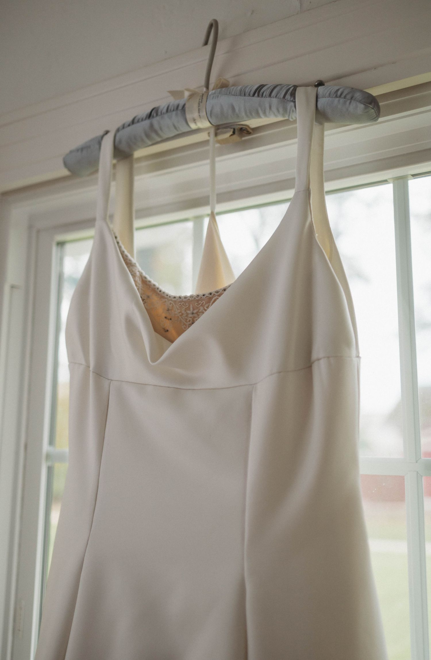 A white formal dress with thin straps hangs on a padded hanger by a sunlit window.