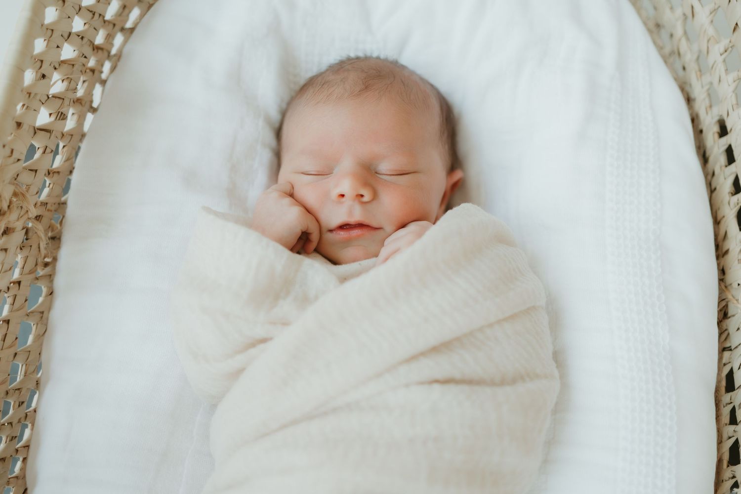 A sleeping newborn baby wrapped in a soft white blanket lies peacefully against a textured backdrop.