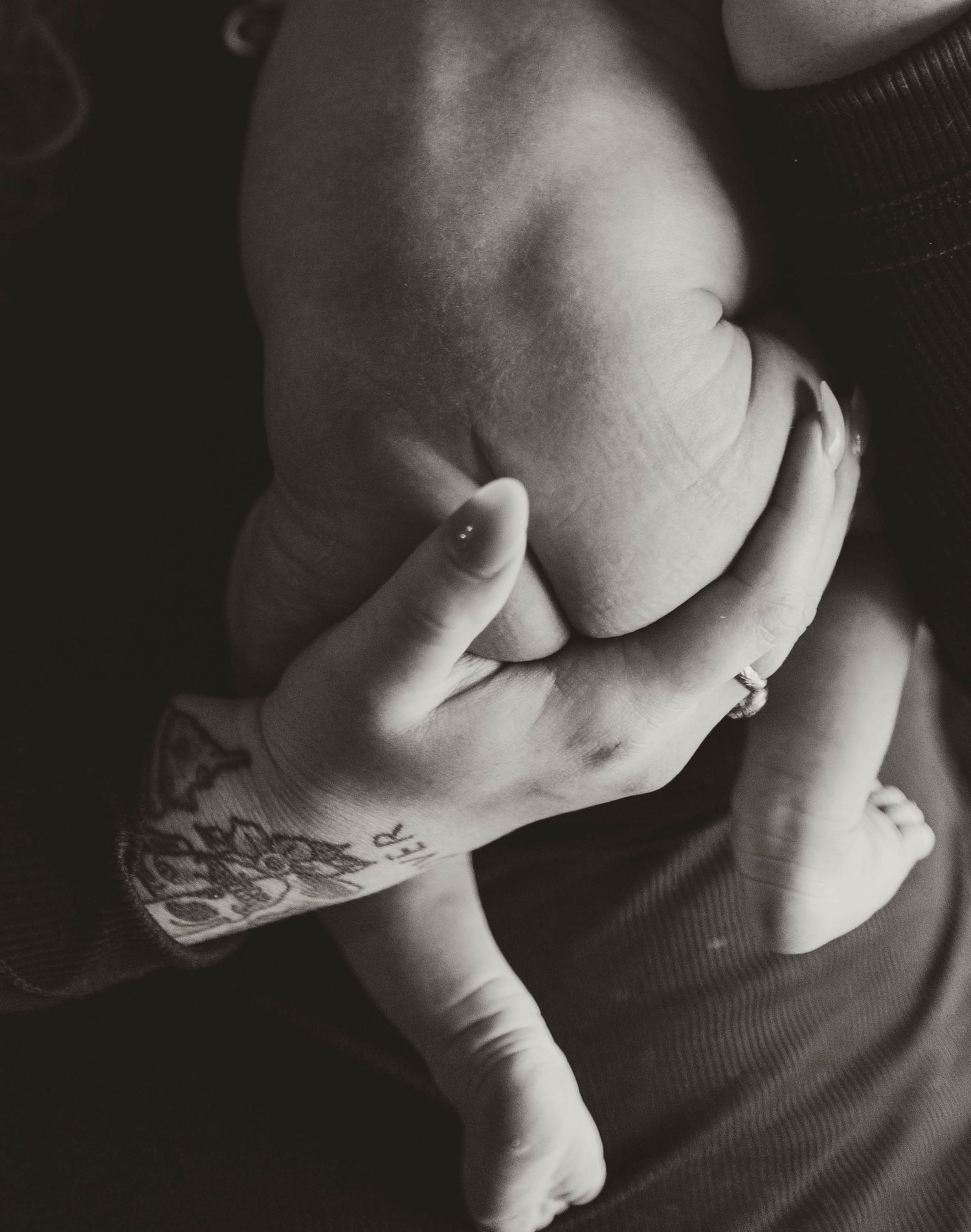 Black and white photograph of hands holding an infant against a dark background, with visible tattoo art on the wrist.