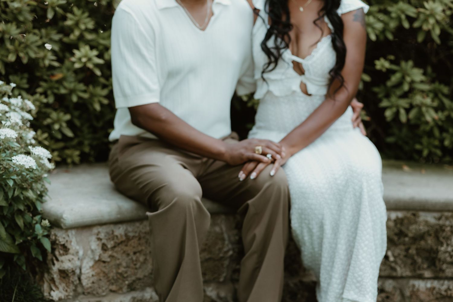 Couple sitting together on a stone bench outdoors, holding hands, dressed in white and tan outfits.