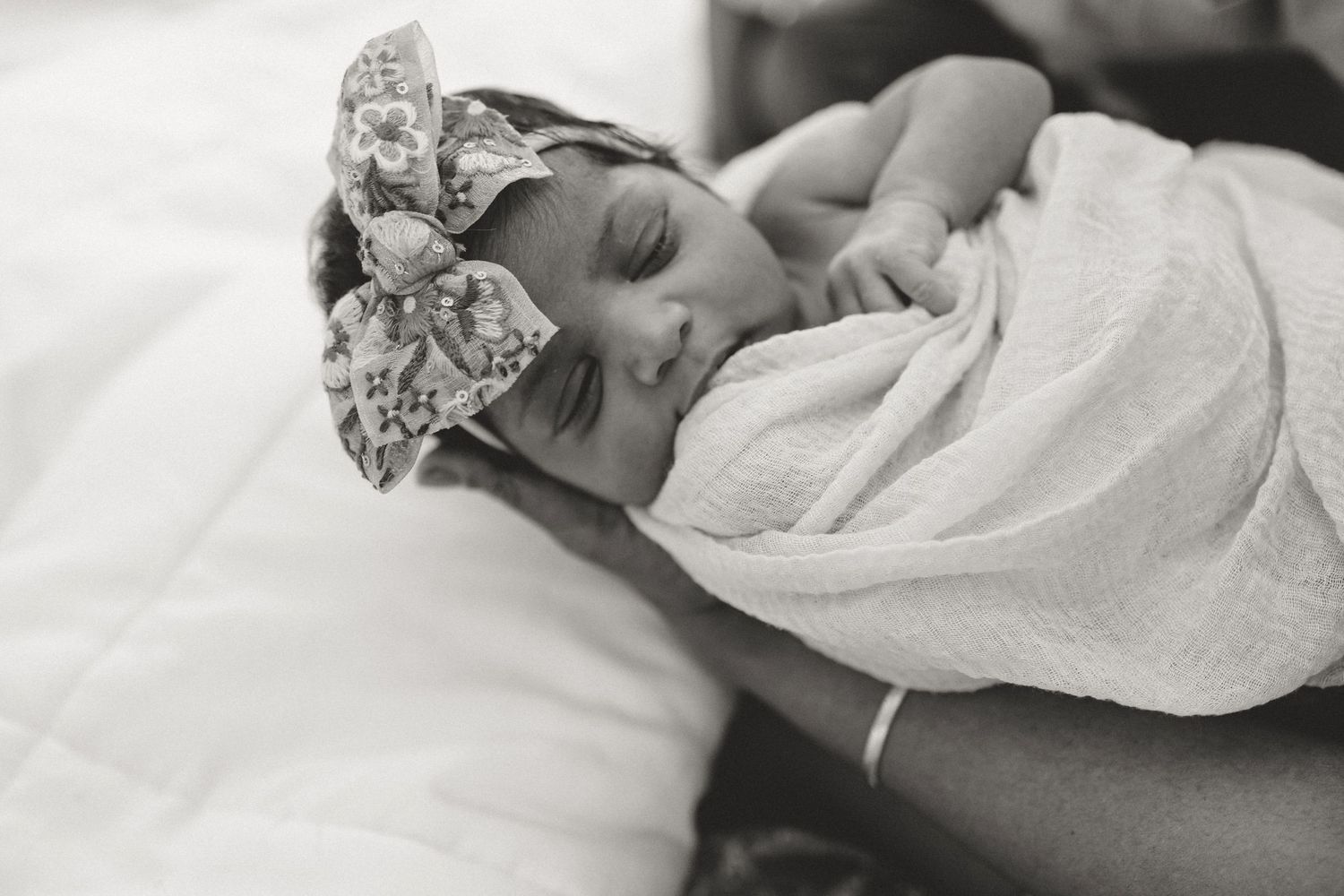 Black and white photo of a sleeping newborn baby wrapped in white swaddle wearing a floral headband.