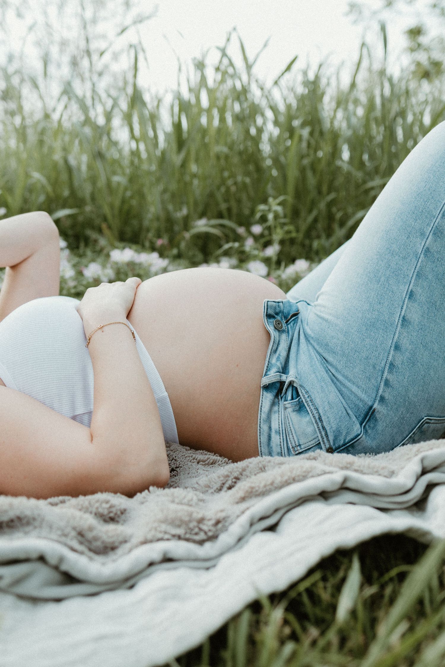 Pregnant woman lying on a blanket outdoors in grass, wearing white bra and open jeans, showing baby bump.