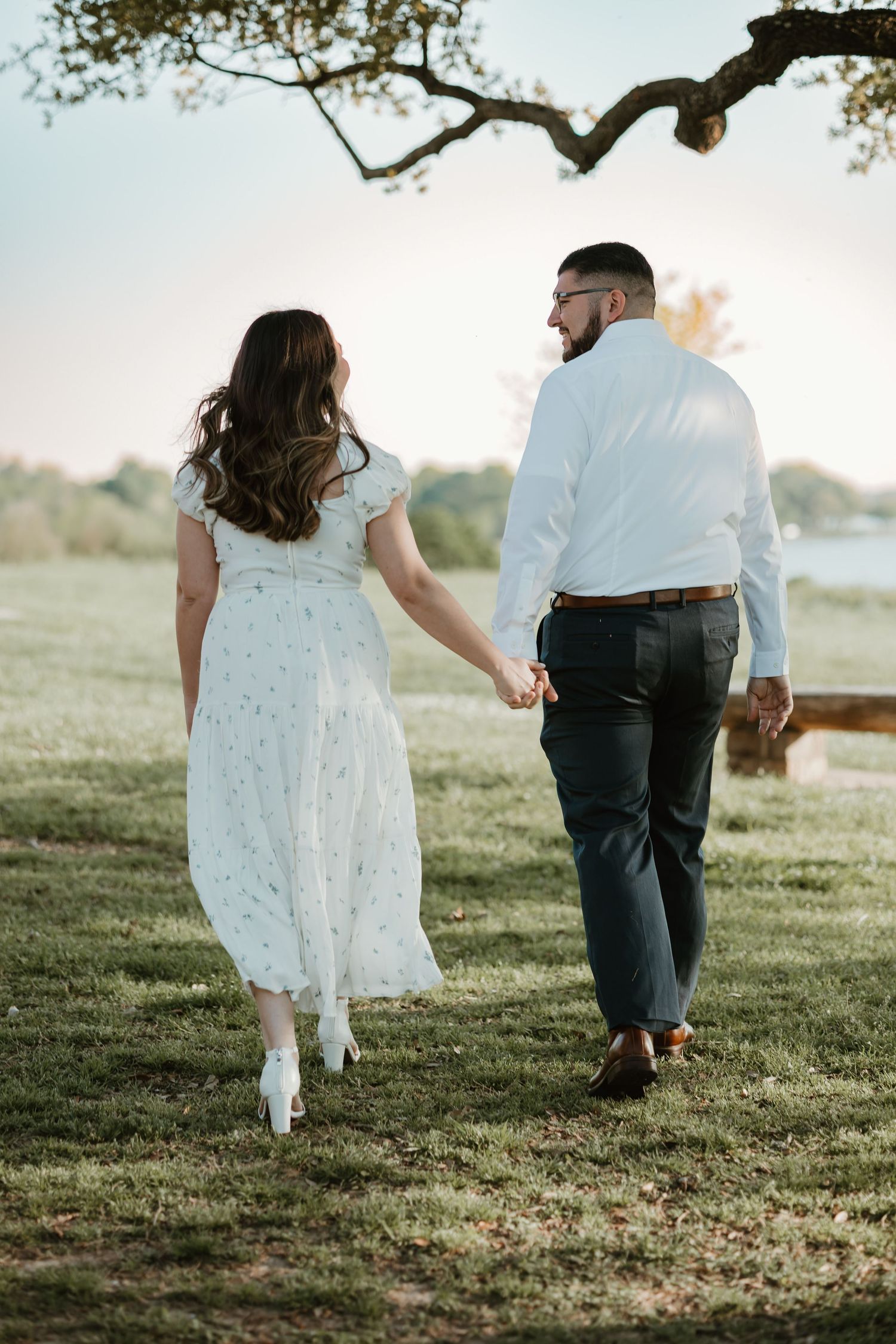 A couple holding hands while walking on grass under a tree branch during a romantic outdoor photoshoot.