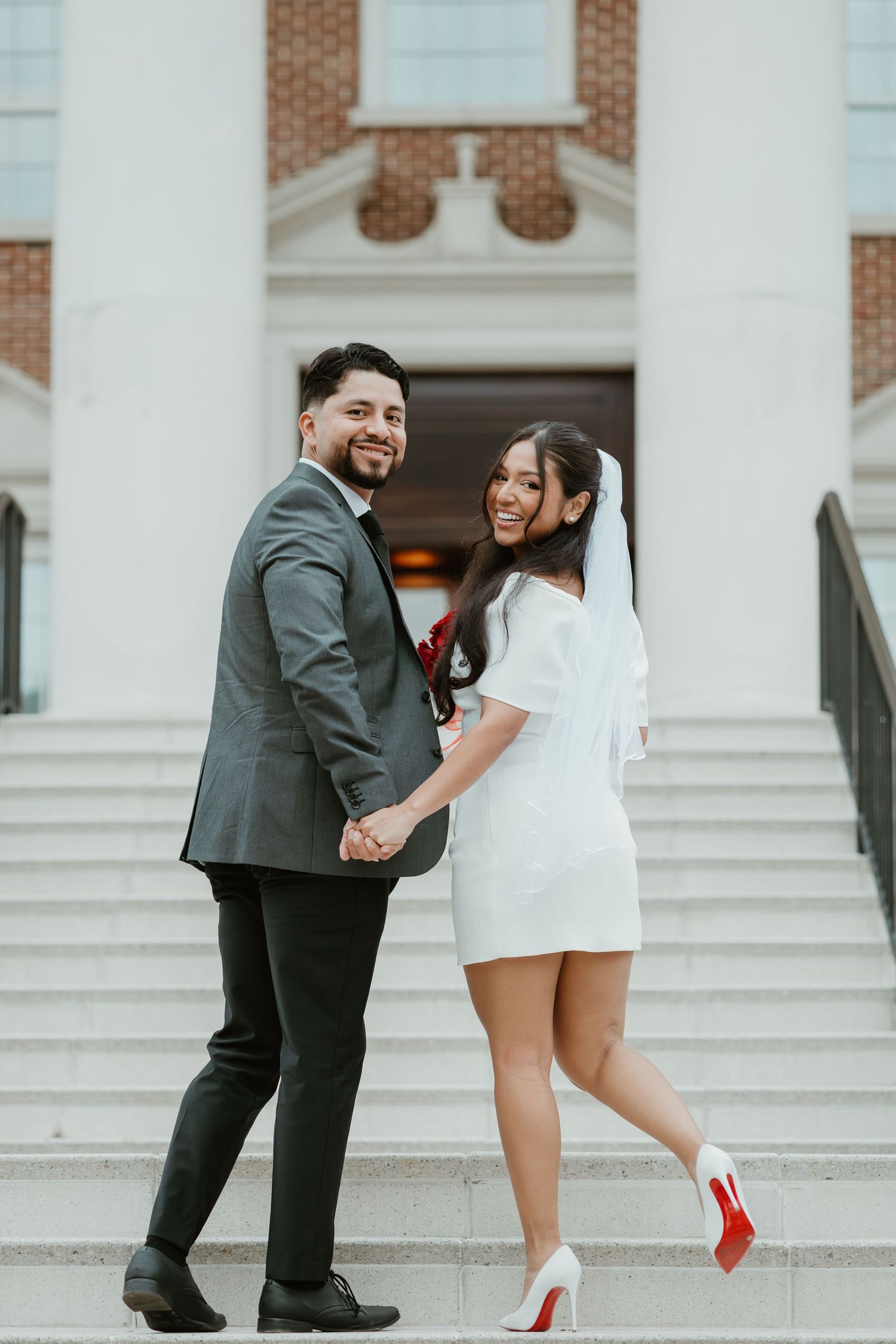 Happy newlywed couple holding hands on church steps, bride in short white dress and veil with red Louboutins, groom in gray suit.