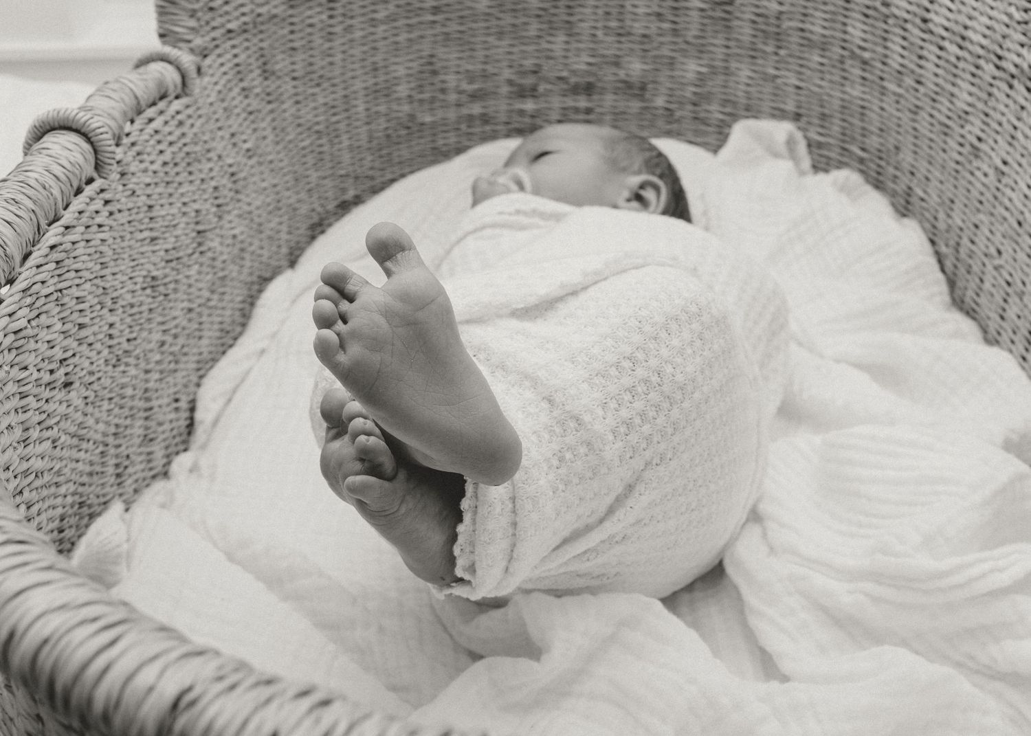 Black and white photo of a sleeping newborn baby wrapped in white blanket inside a wicker bassinet.
