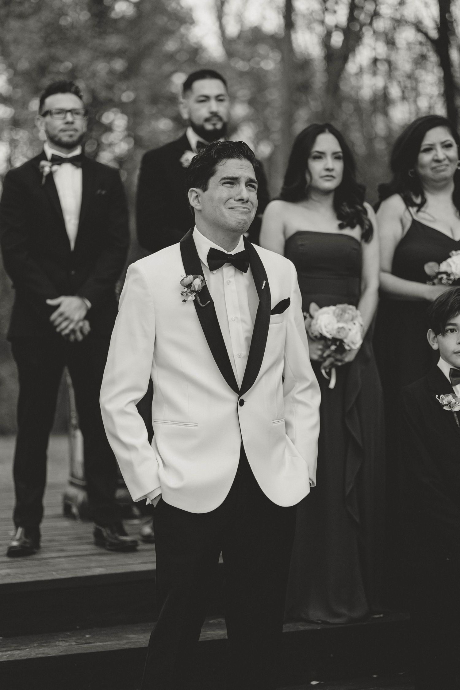 A black and white wedding photo showing a groom in white tuxedo jacket posed with bridesmaids in dark dresses.