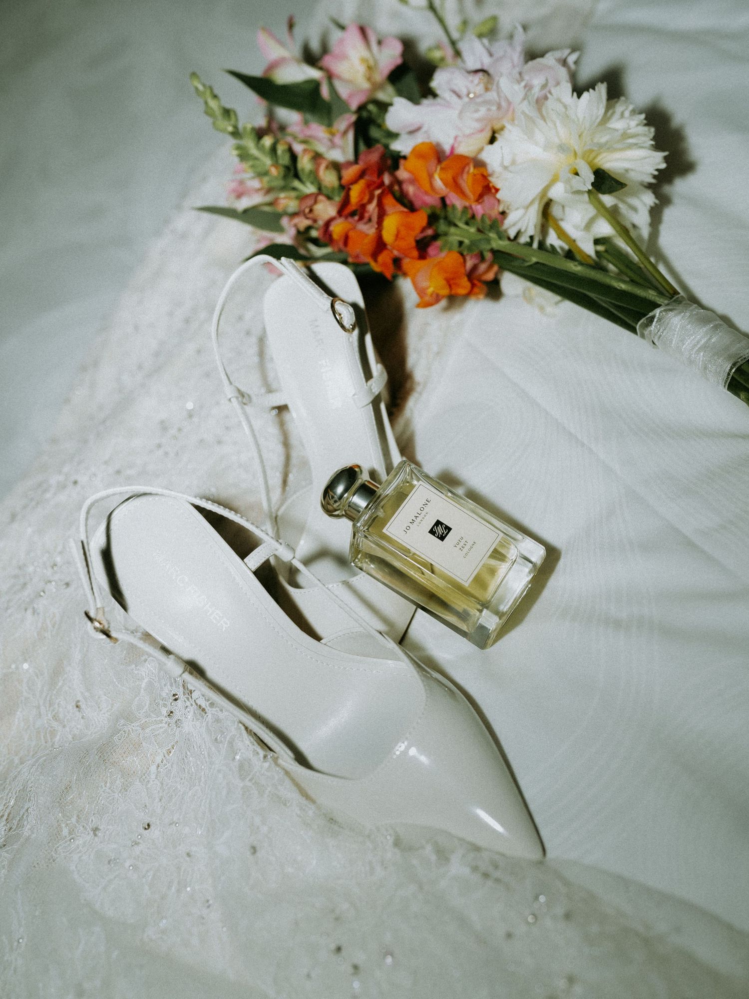 White bridal heels and Jo Malone perfume resting on a wedding dress beside a colorful floral bouquet.