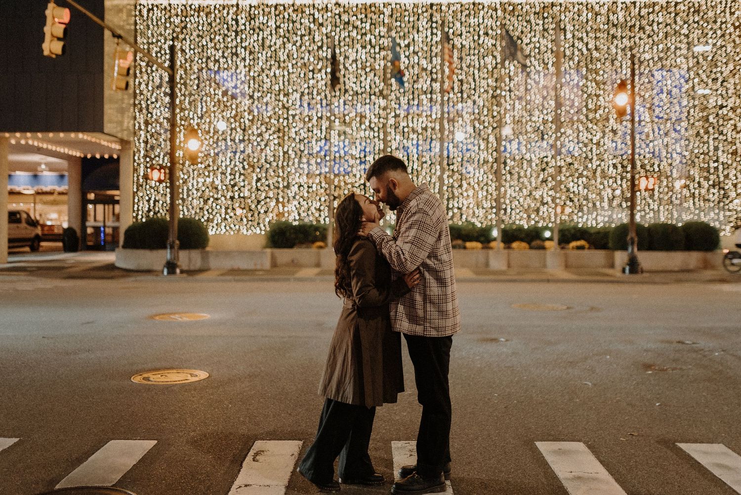 A couple shares an intimate moment on a city crosswalk at night with glowing holiday lights strung overhead.