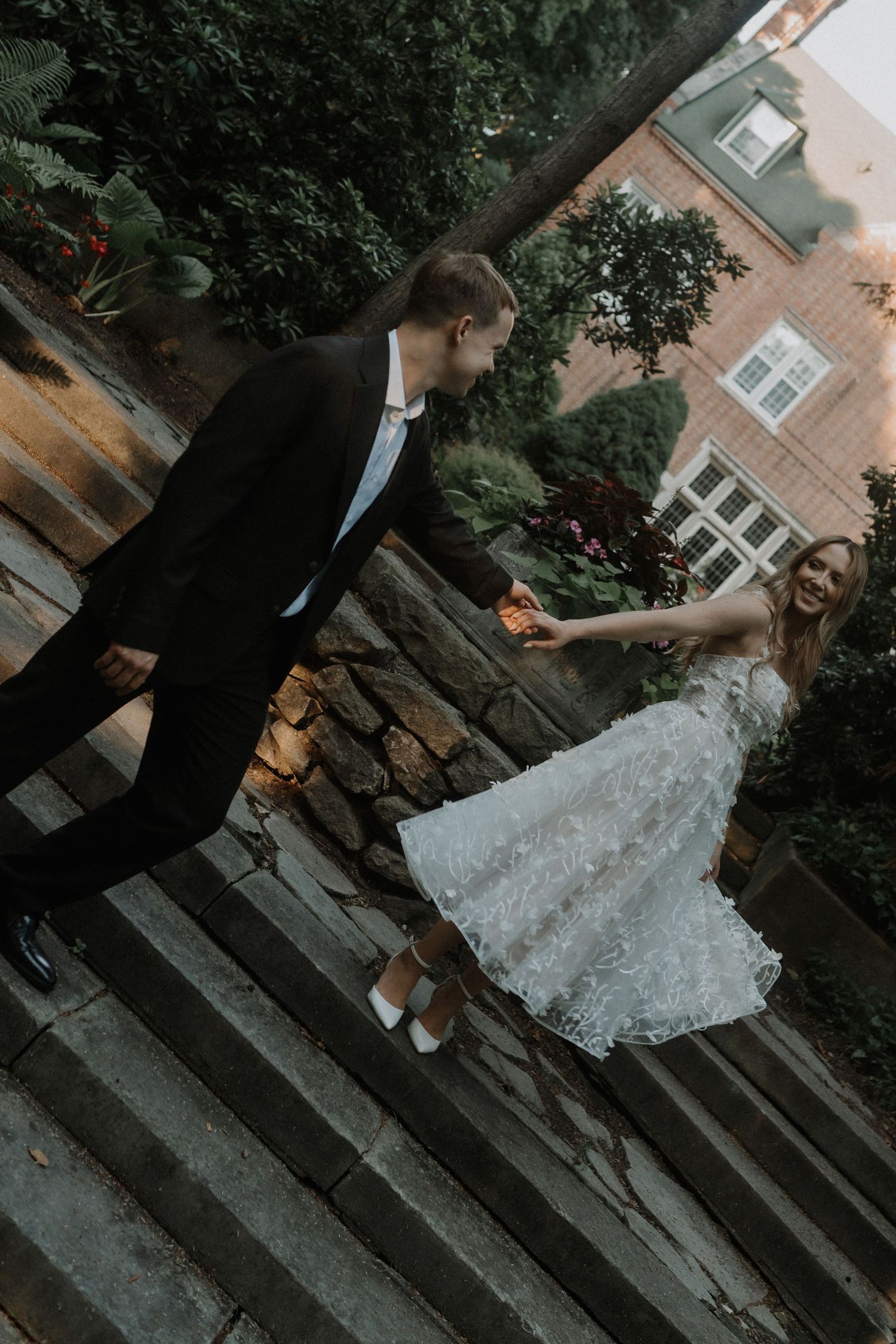 A couple in formal wedding attire walk down outdoor stone steps in a garden setting at dusk.