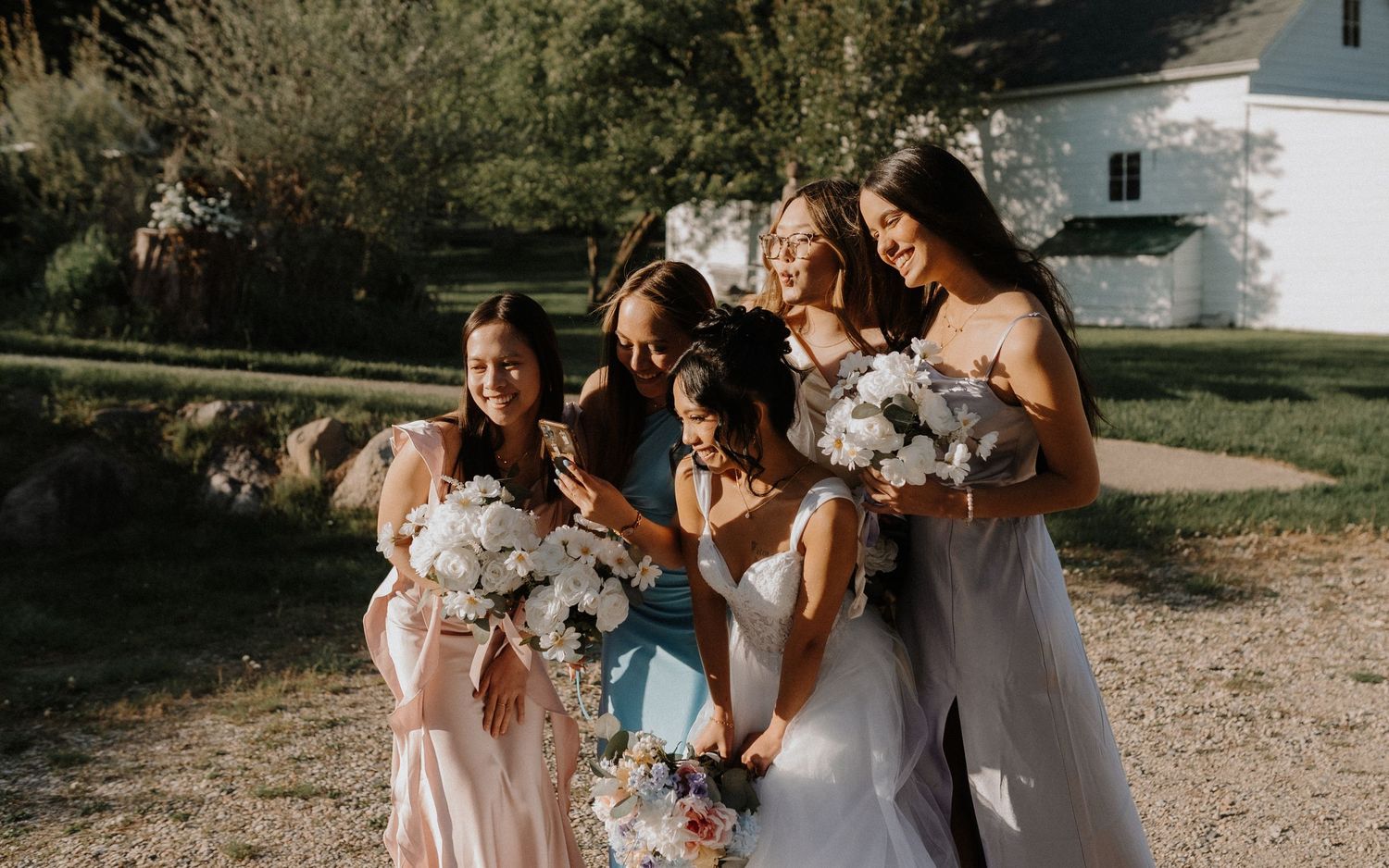 A group of bridesmaids in pastel dresses gather together while holding white floral bouquets at an outdoor wedding.