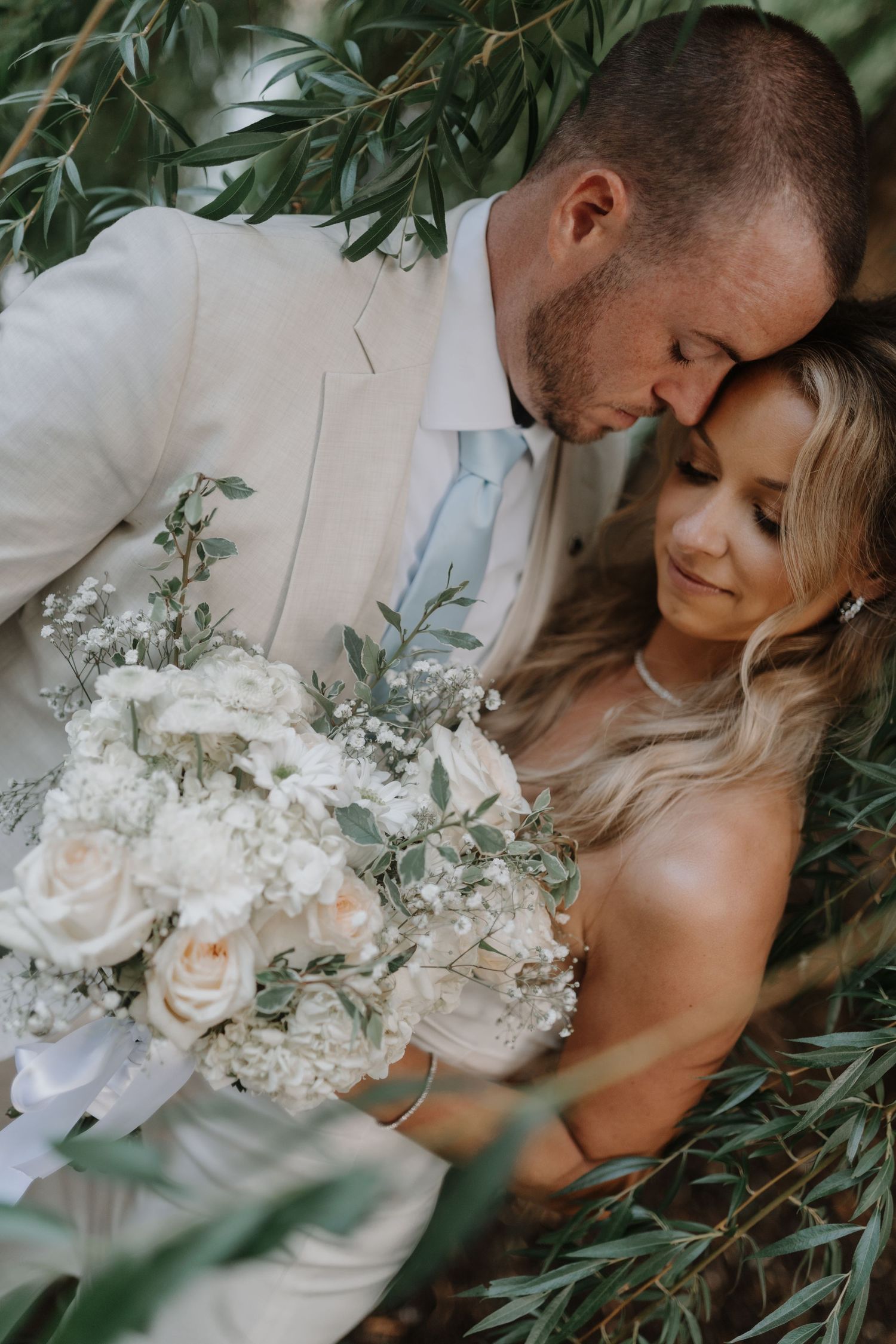 Romantic wedding couple embraces among greenery with white and peach floral bouquet in a series of intimate moments.