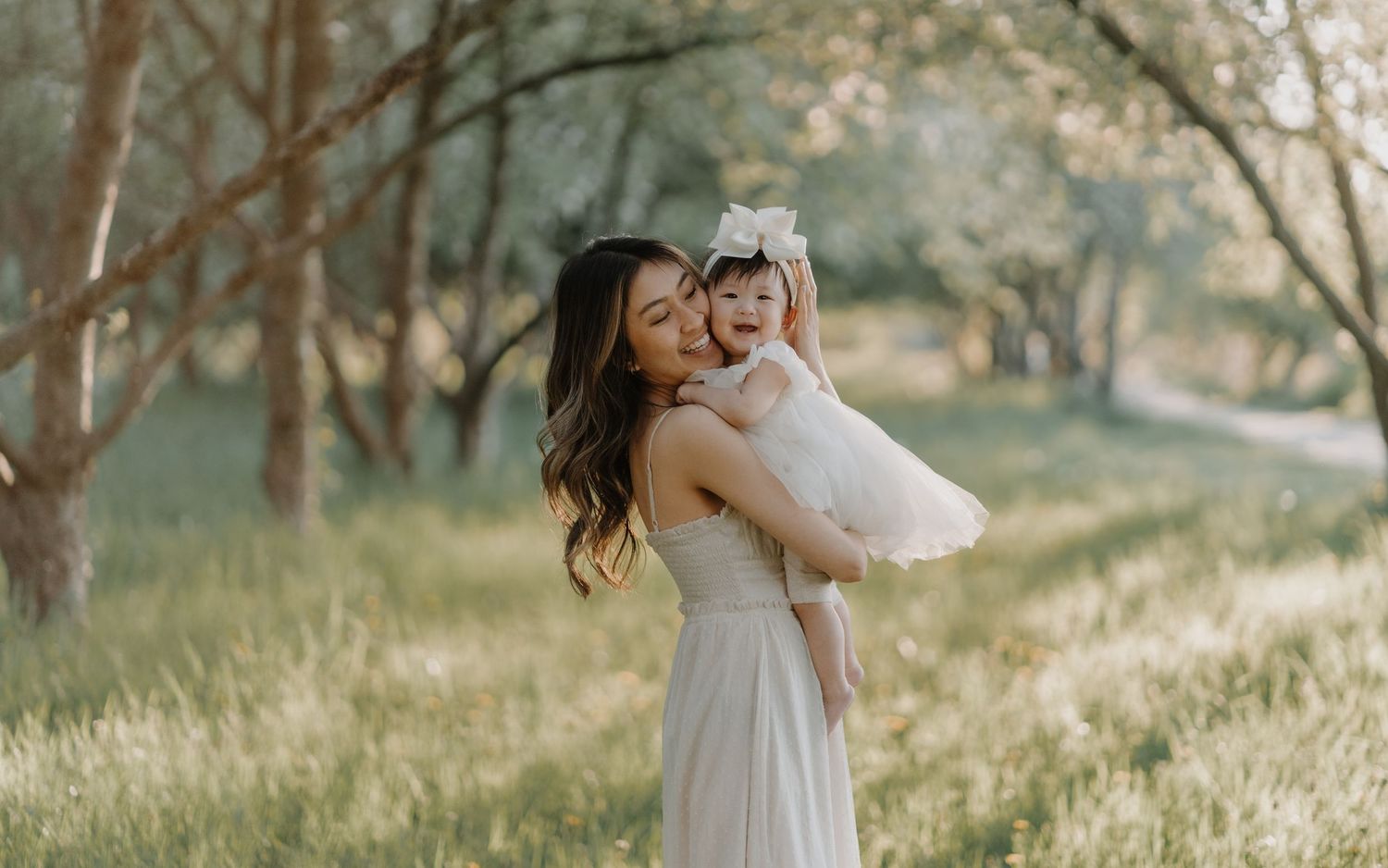 A parent and child wearing white dresses embrace outdoors in a natural setting with sunlit grass and trees.