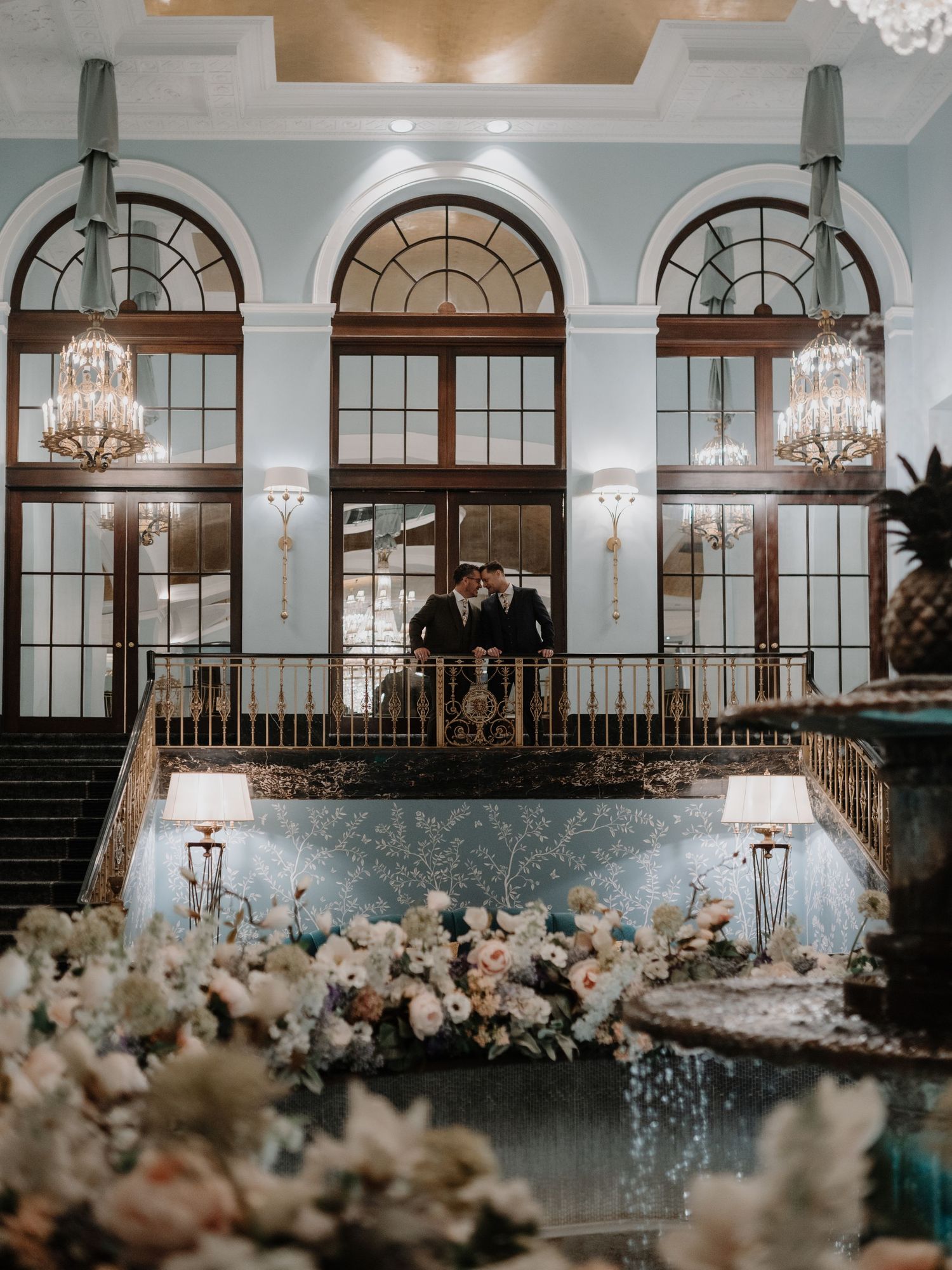 Two grooms share a romantic moment on a grand balcony adorned with crystal chandeliers and lush floral arrangements.