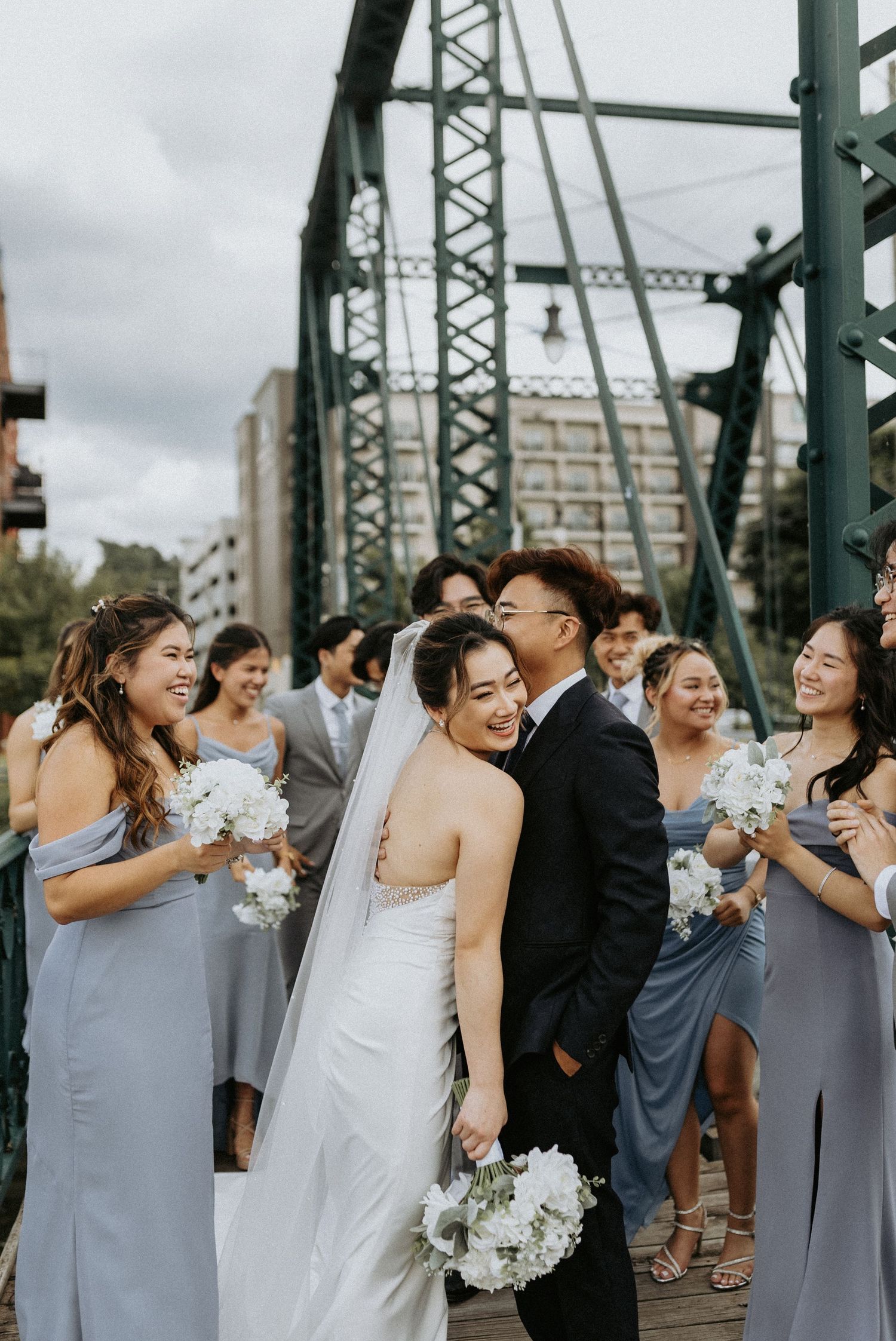 Wedding party in gray and white attire shares joyful laughter on an iron bridge during outdoor photo session.