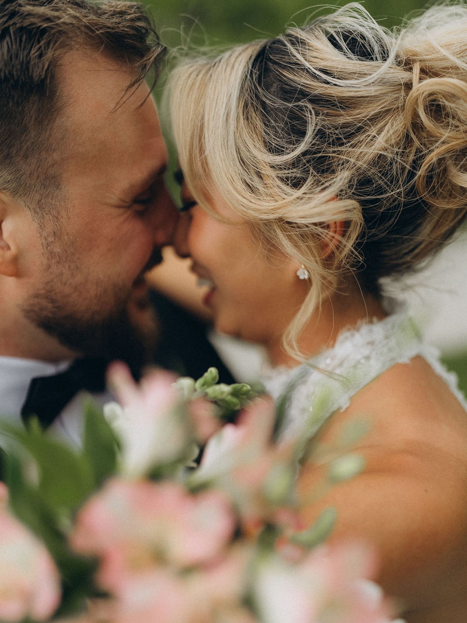 A bride and groom share a romantic moment, nearly kissing, with soft pink wedding flowers in the foreground.