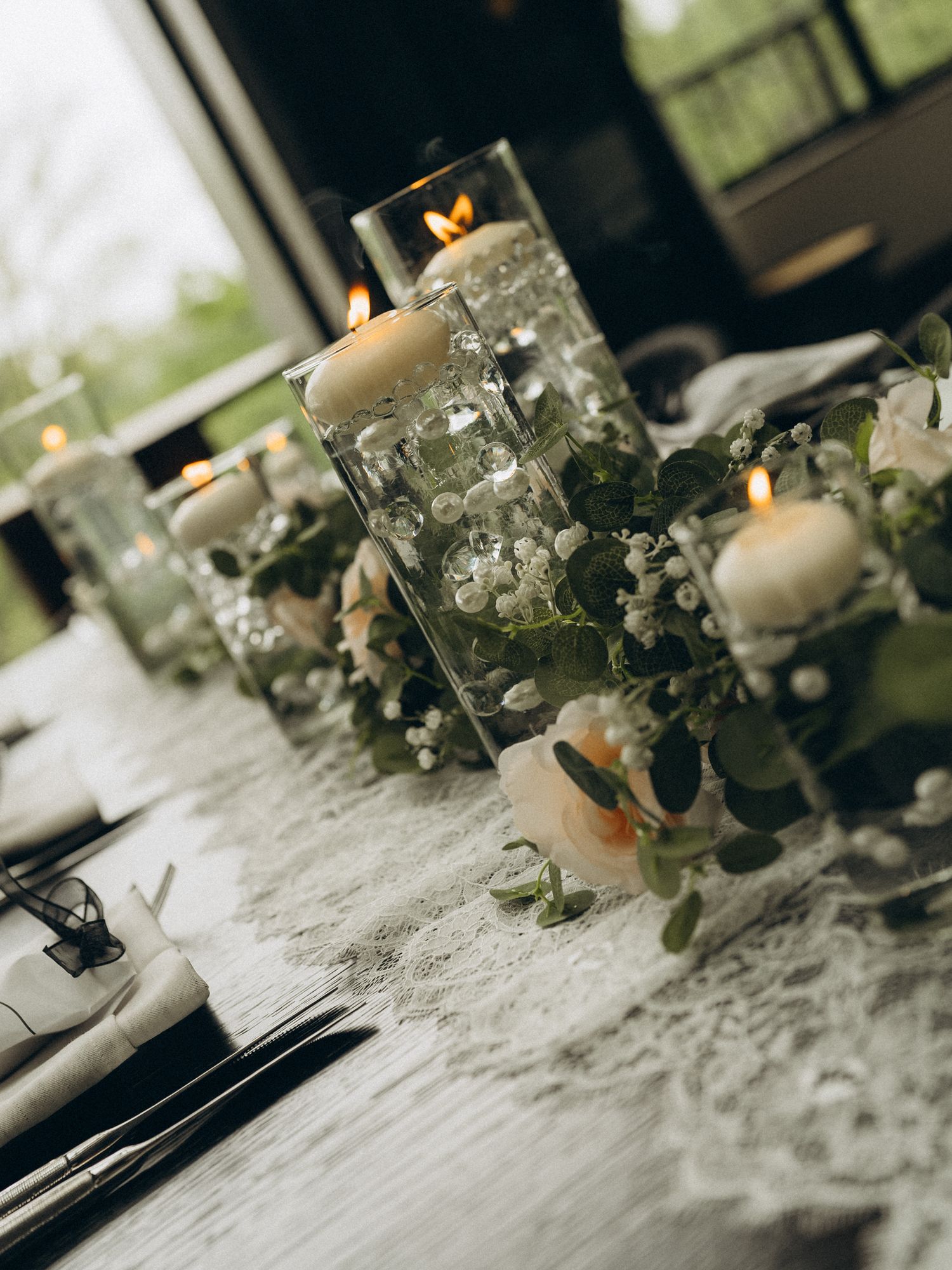 Elegant wedding table centerpiece with floating candles in glass cylinders, pearl beads, and soft pink roses with greenery.