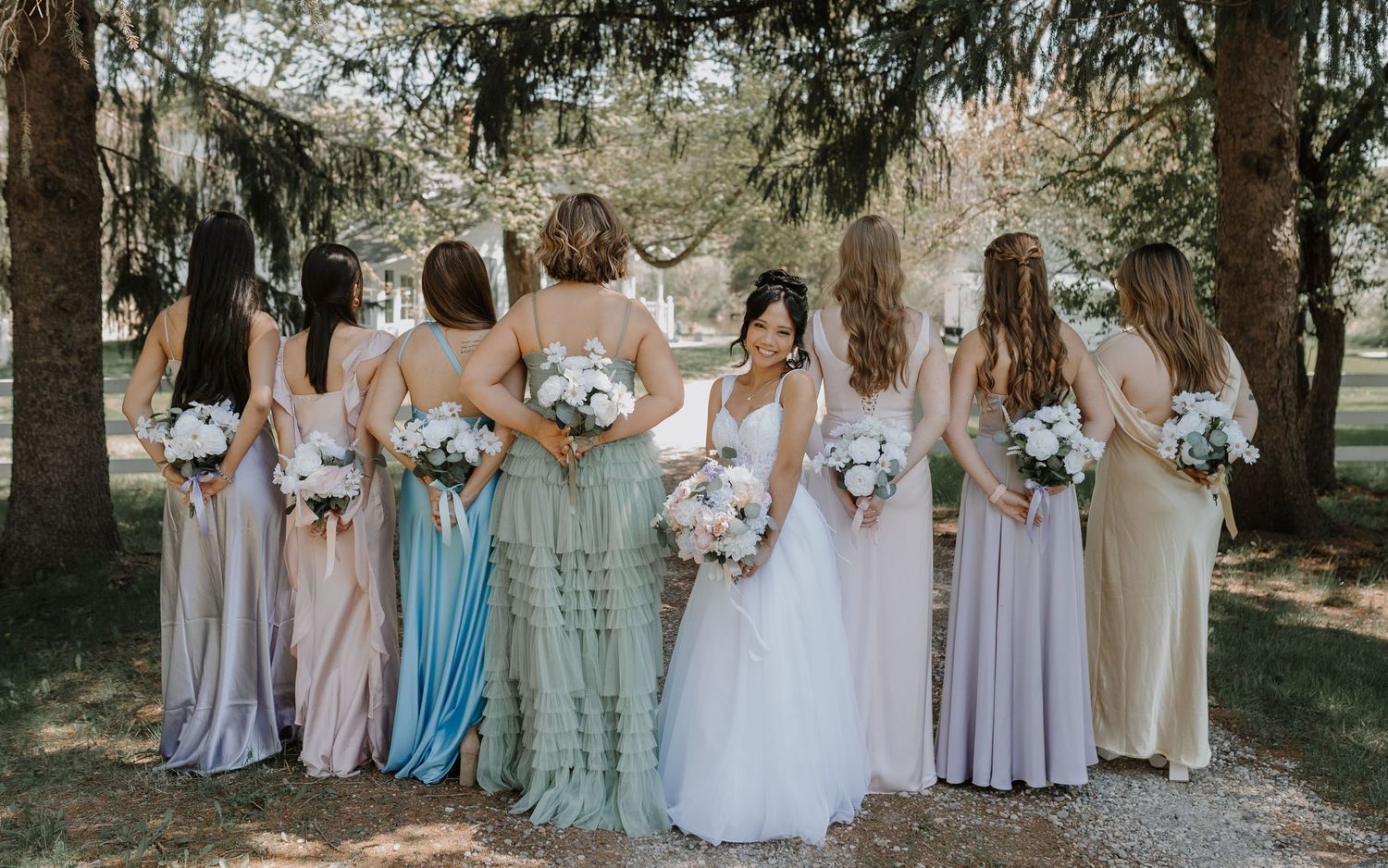 Bridesmaids in soft pastel lavender and mint dresses stand in a line holding floral bouquets during an outdoor wedding.