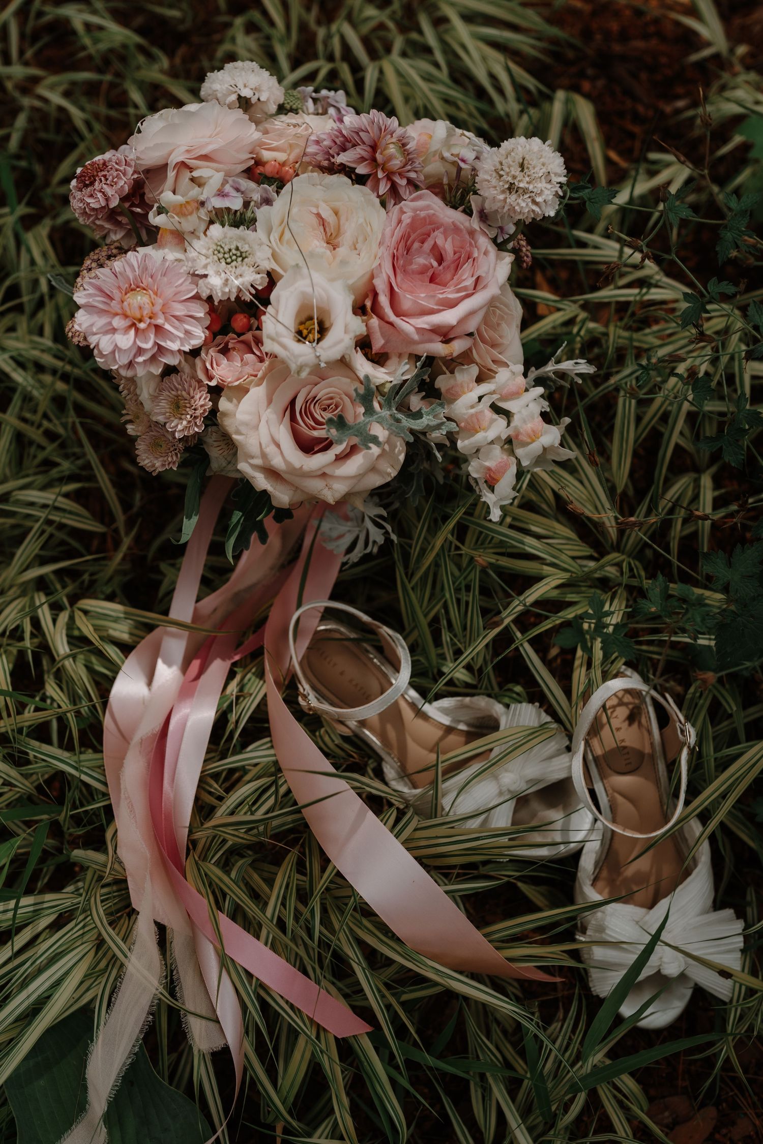A romantic bridal bouquet with pink roses and mixed flowers with matching ribbon lies beside ballet slippers on grass.