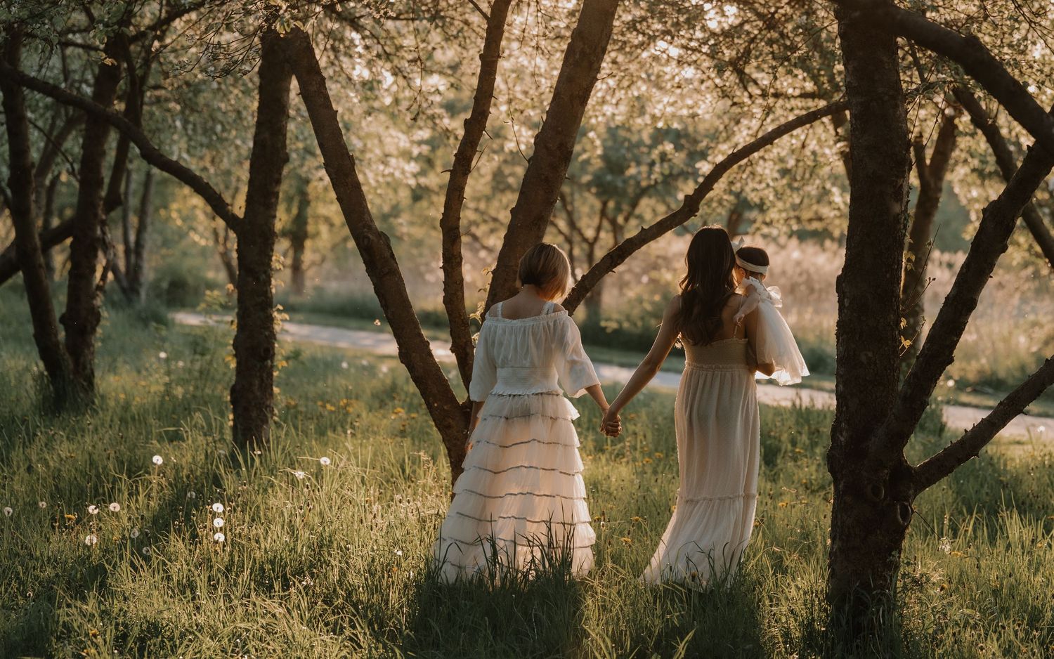 Two people in flowing white dresses walk together through a sunlit forest grove on a dreamy summer afternoon.