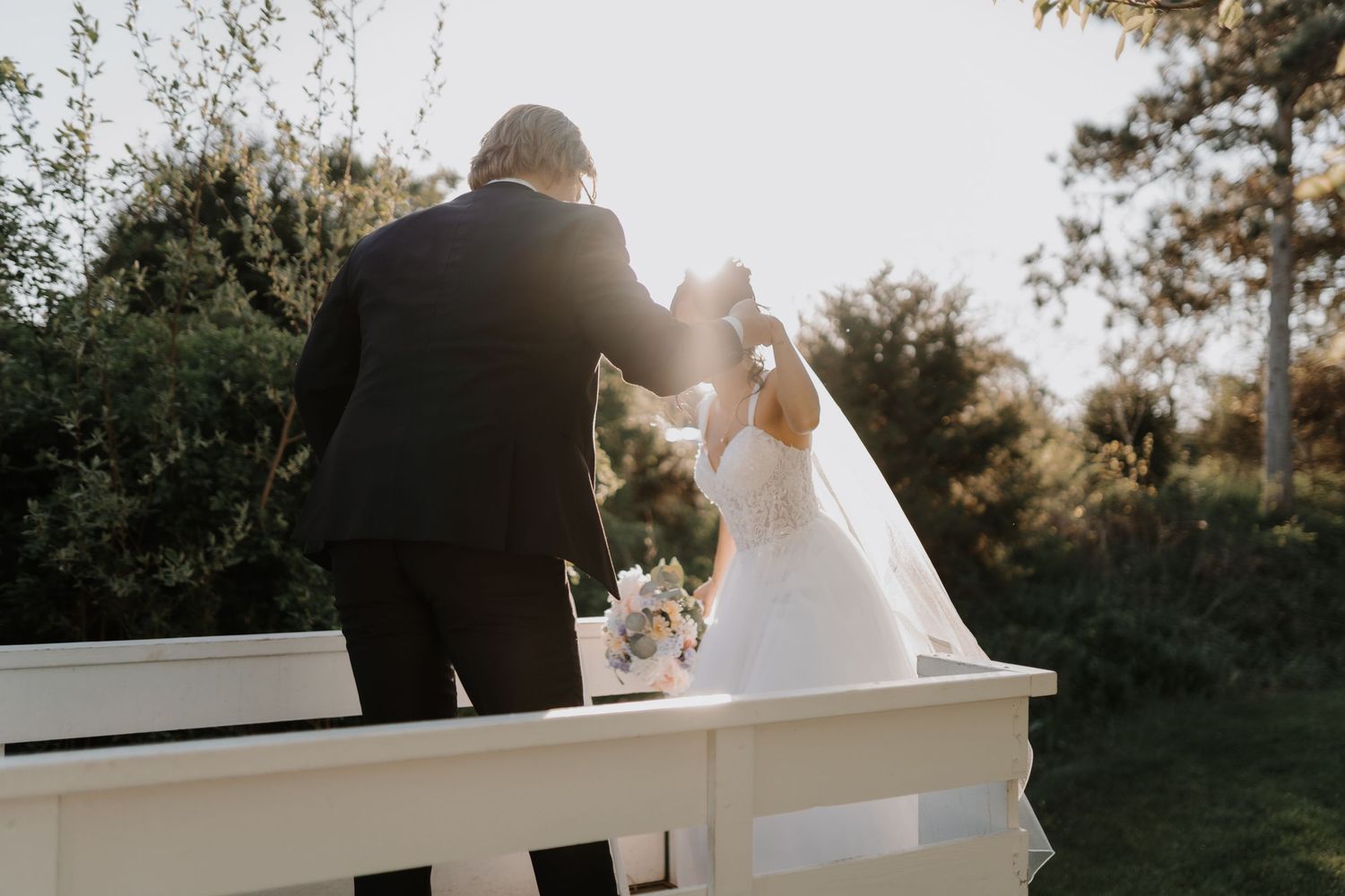 A couple in wedding attire stand together on a white balcony during sunset with trees in the background.