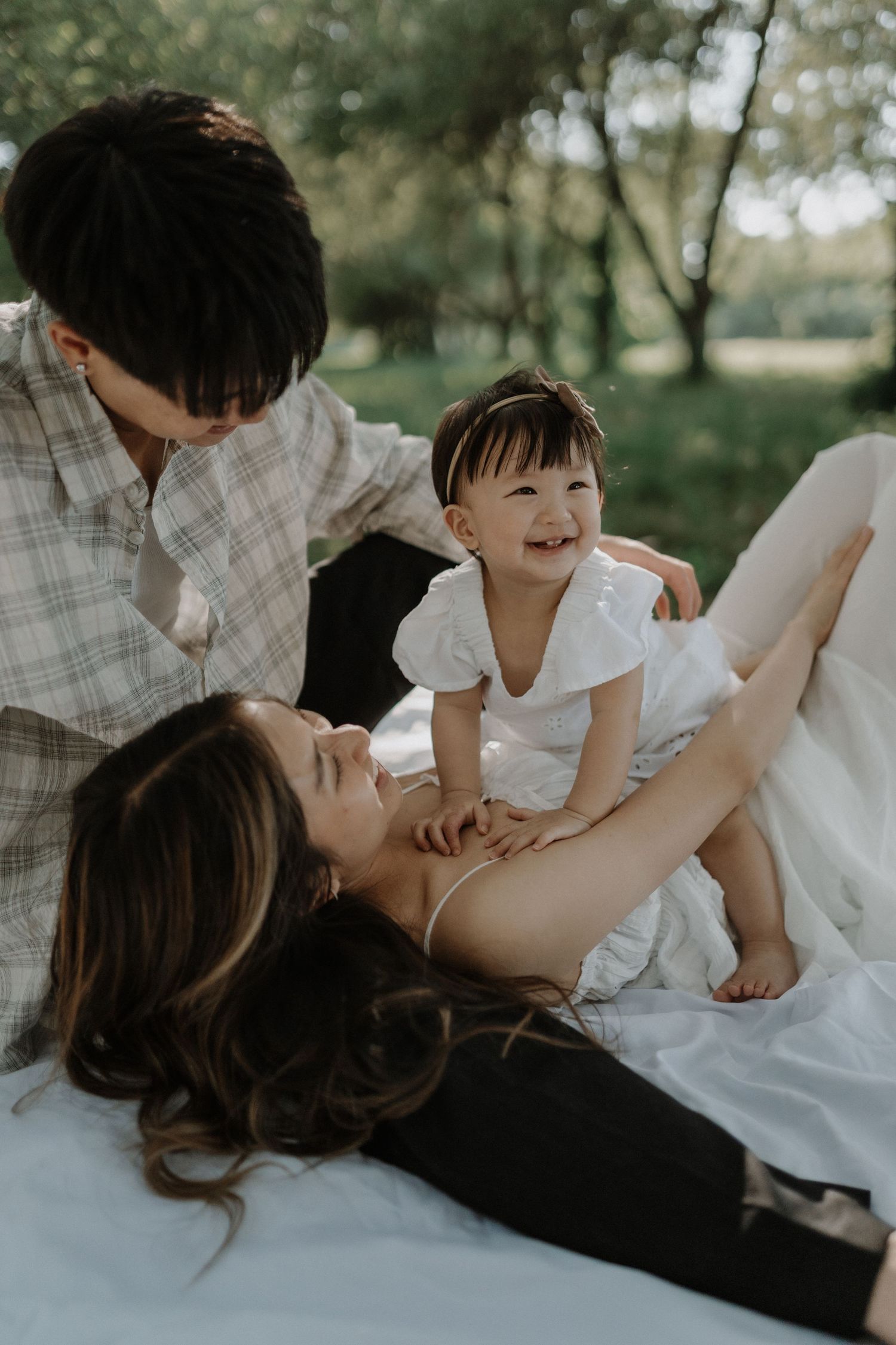 A tender family moment captured outdoors showing playful interaction and laughter while laying in white clothing on grass.