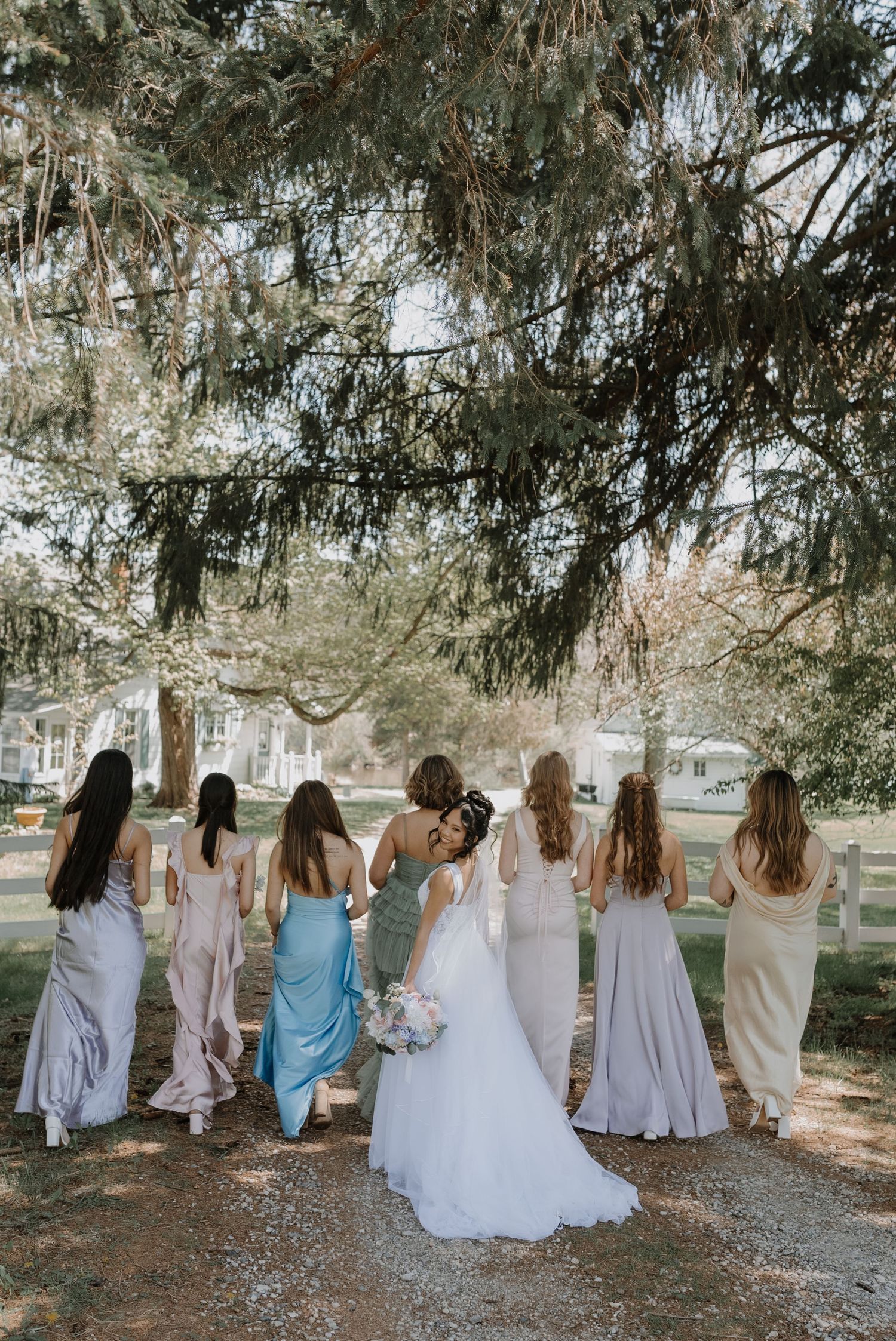 A bridal party wearing pastel dresses walking together under swaying trees on a sunny afternoon.