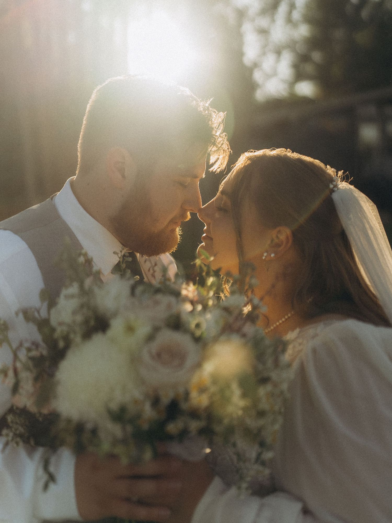 A bride and groom share a romantic moment, foreheads touching, backlit by golden sunlight with a white floral bouquet.