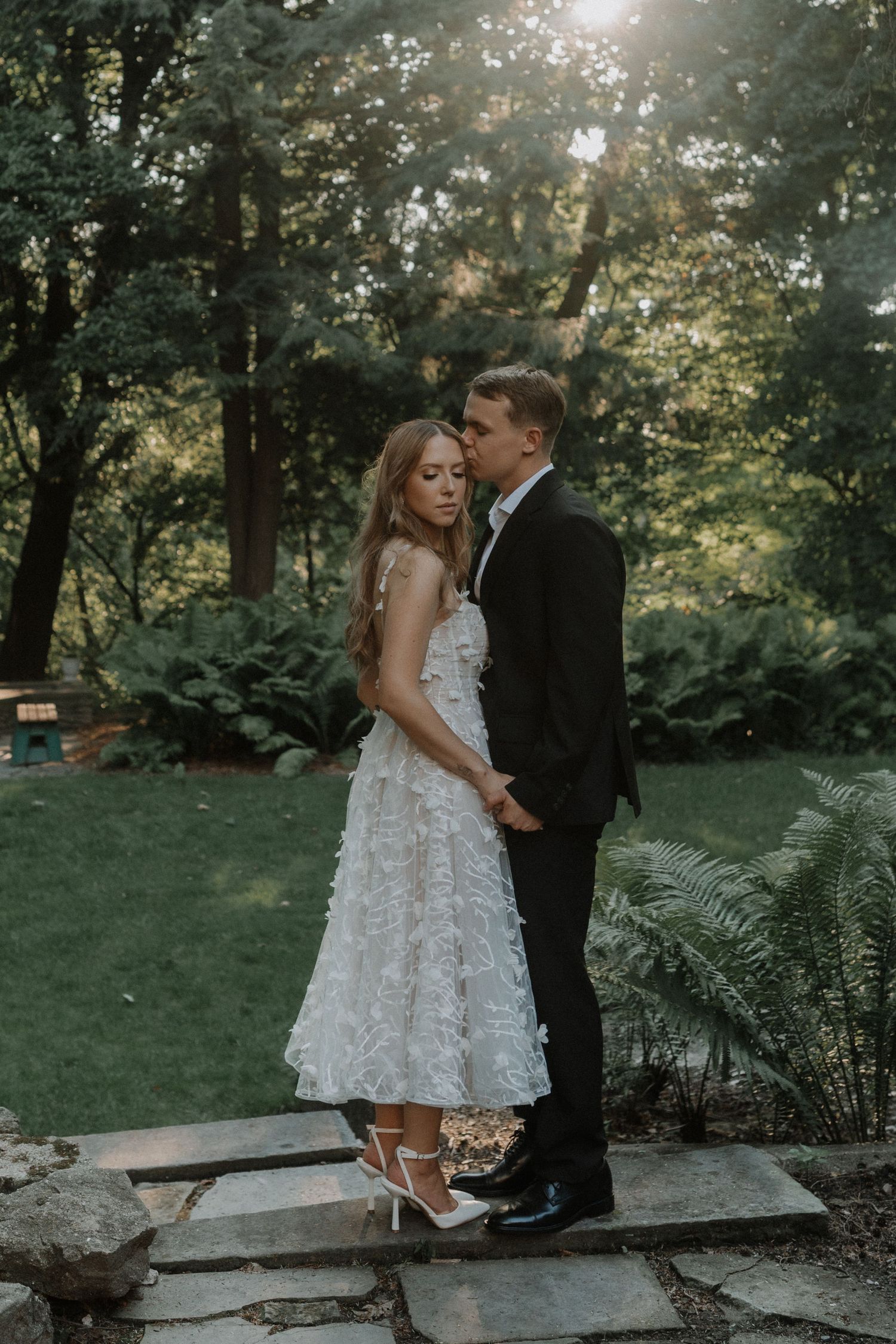 A couple in formal wedding attire embrace on stone steps in a lush garden setting with sunlight filtering through trees.