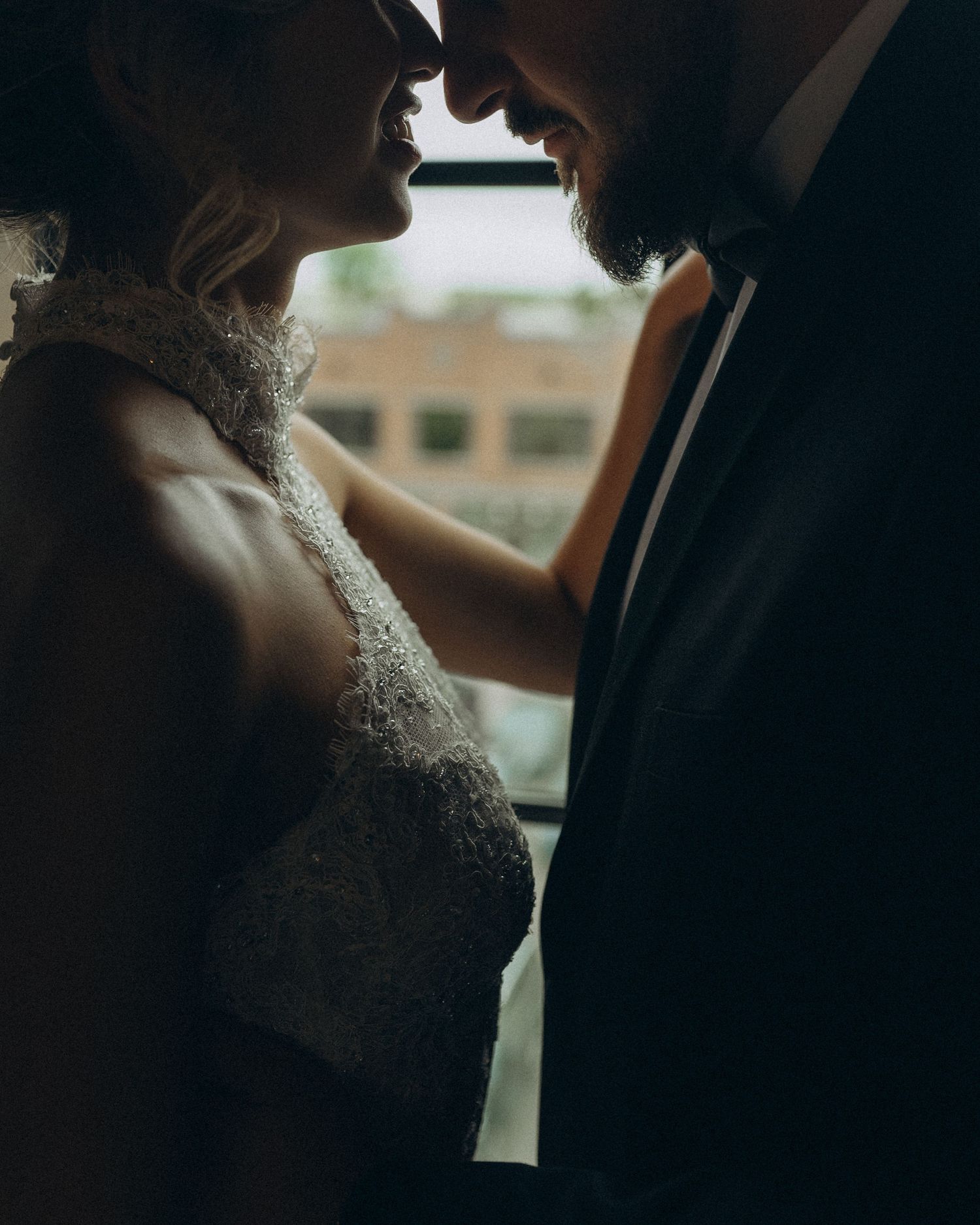 Romantic bride and groom sharing an intimate moment by a window in wedding attire, soft moody lighting.