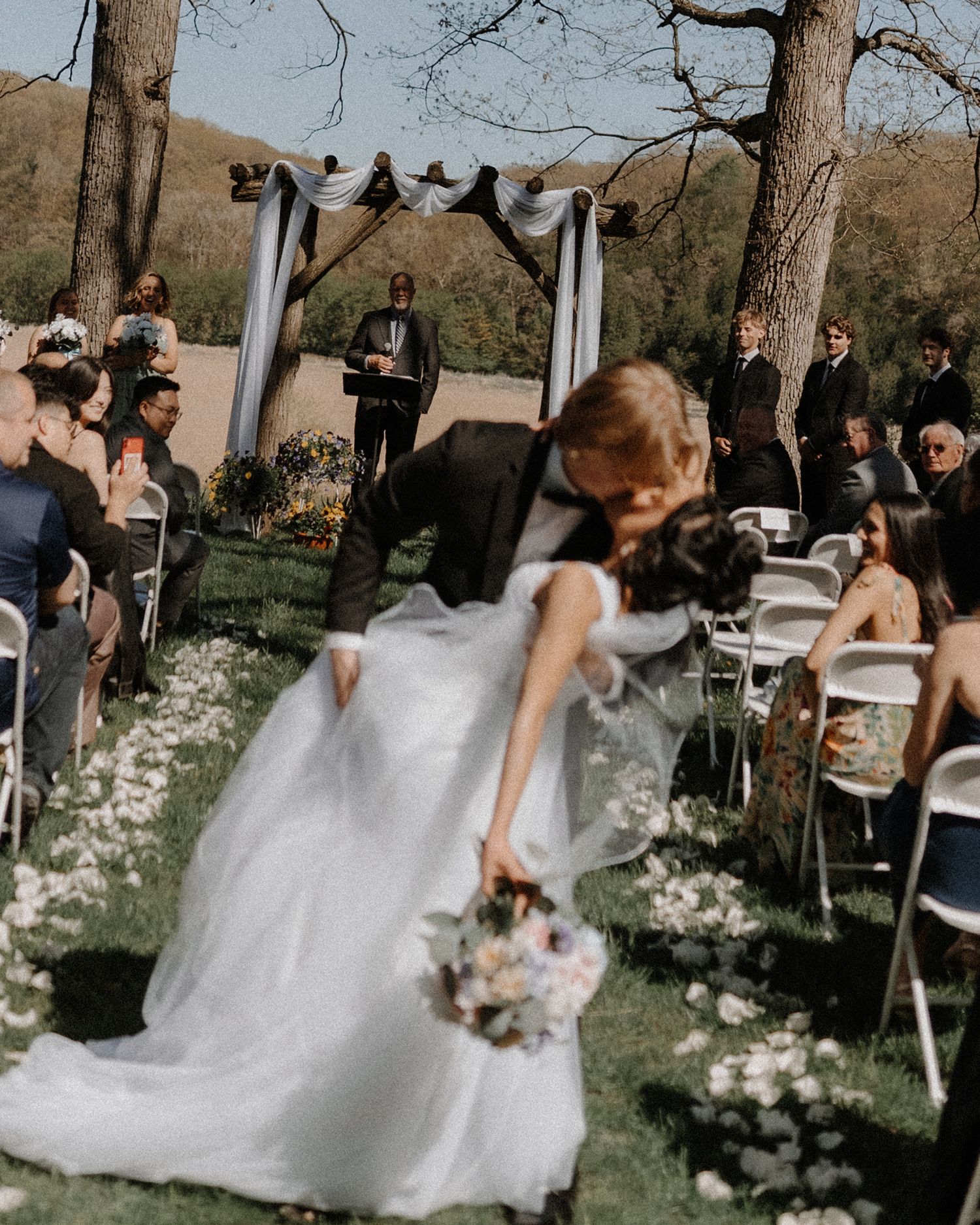 A bride in a white ballgown twirls gracefully during a garden wedding ceremony under tall trees and white floral decor.