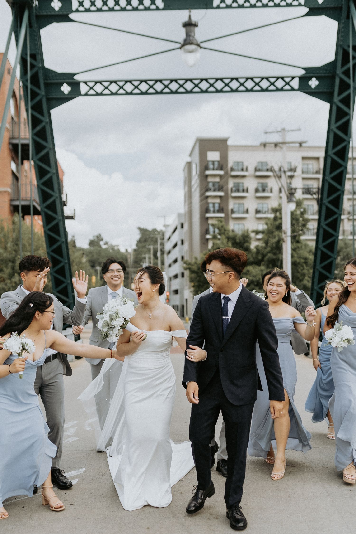 A wedding party walks together and celebrates on an industrial style bridge with grey bridesmaids dresses and white florals.