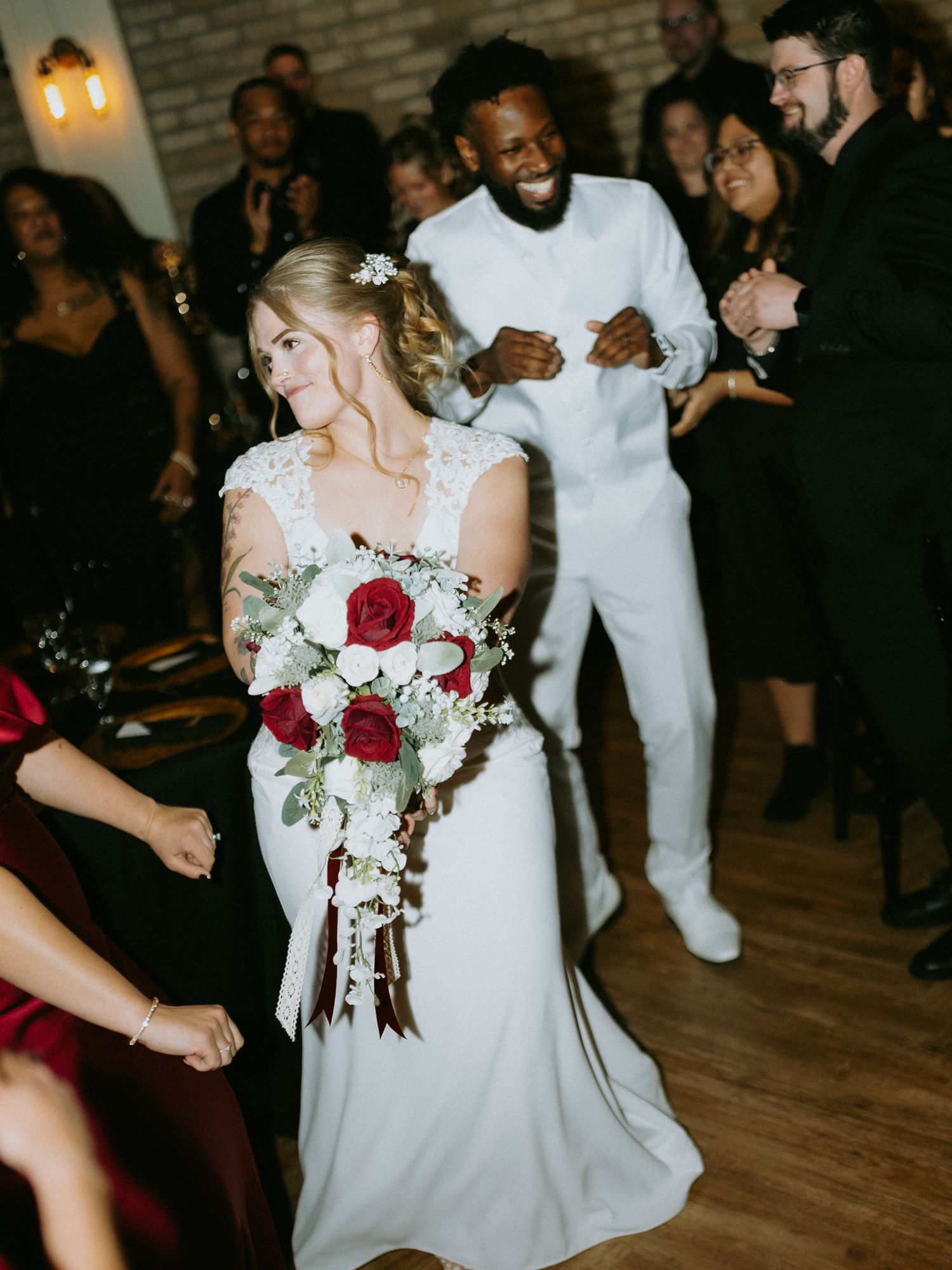 Bride in lace wedding gown holding red rose bouquet dancing with guests at elegant wedding reception.