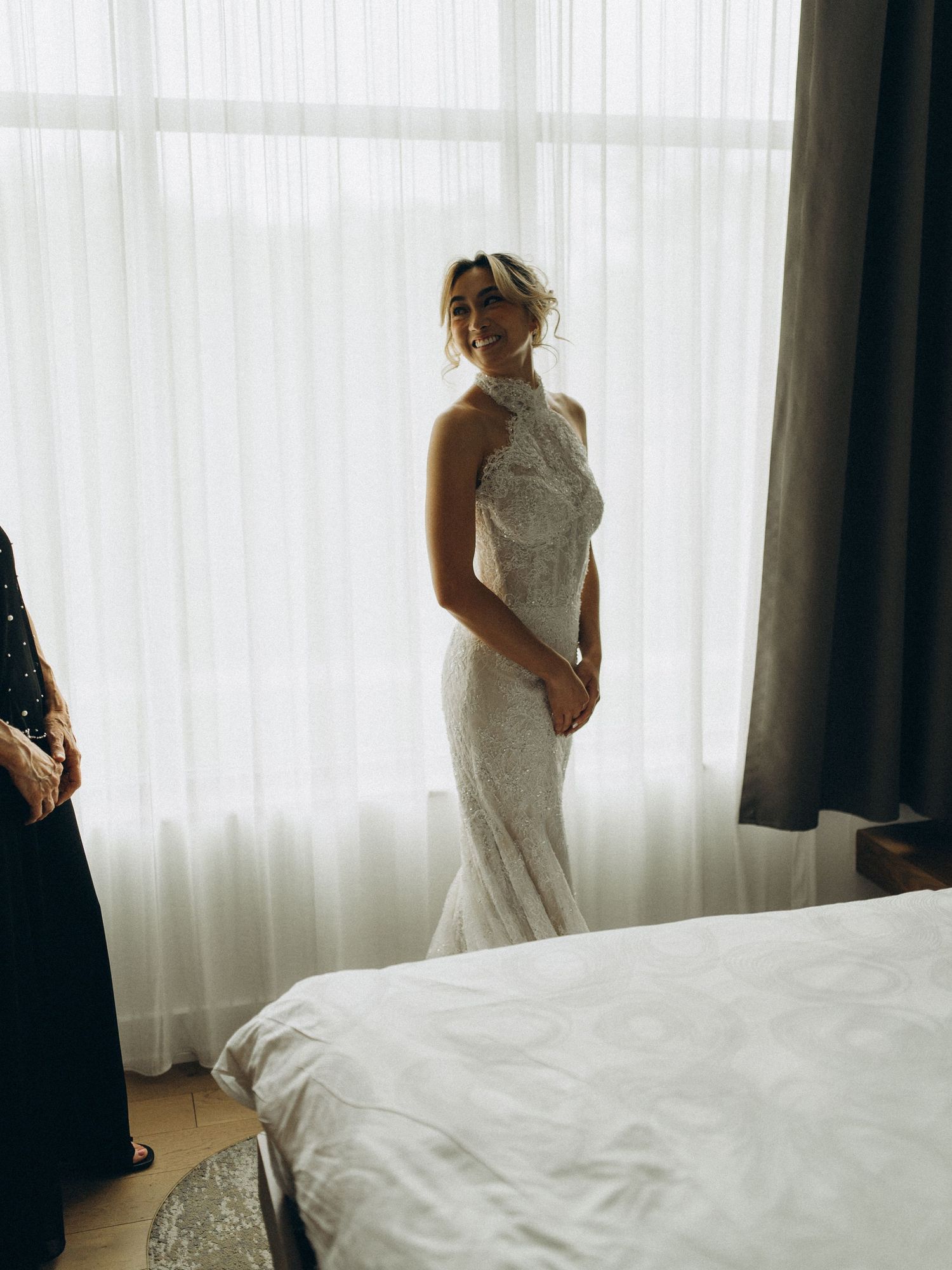 Bride in a beaded halter wedding gown smiling in a sunlit hotel room.