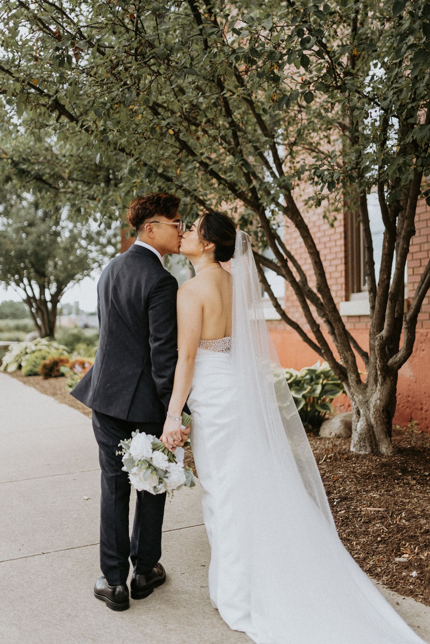 A wedding couple shares a tender moment under spring blossoms, with the bride holding white flowers and wearing a trailing veil.