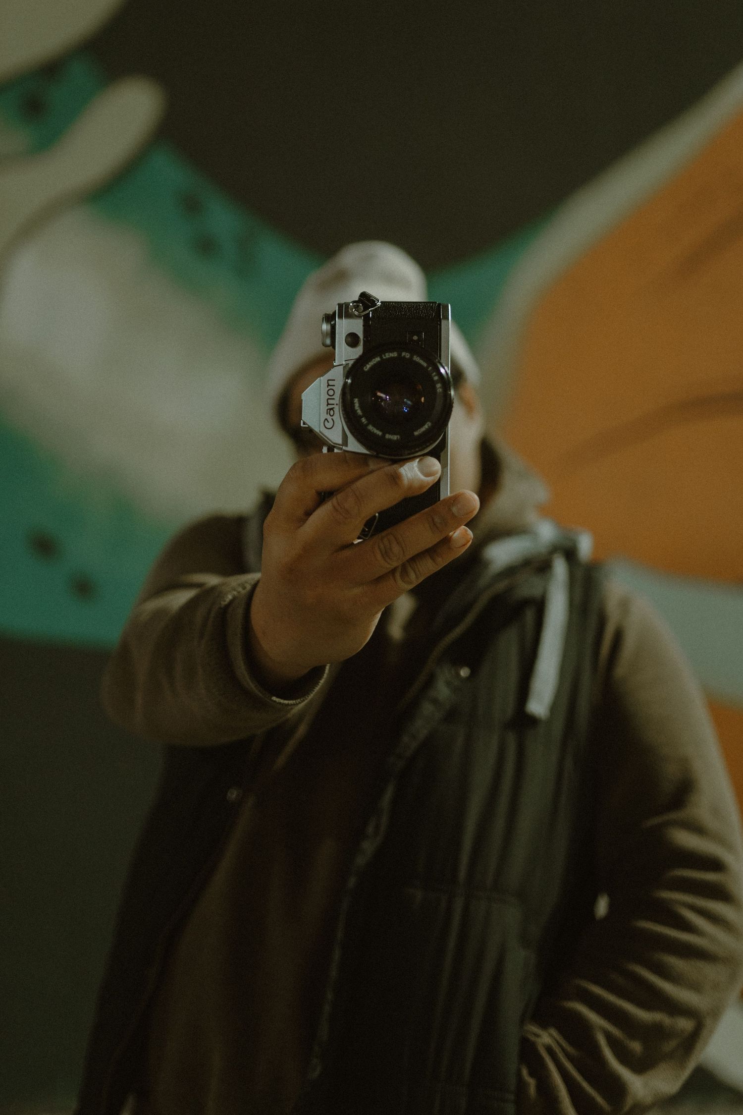 Person in brown jacket and white hat taking a mirror selfie with a vintage film camera in front of colorful graffiti wall.