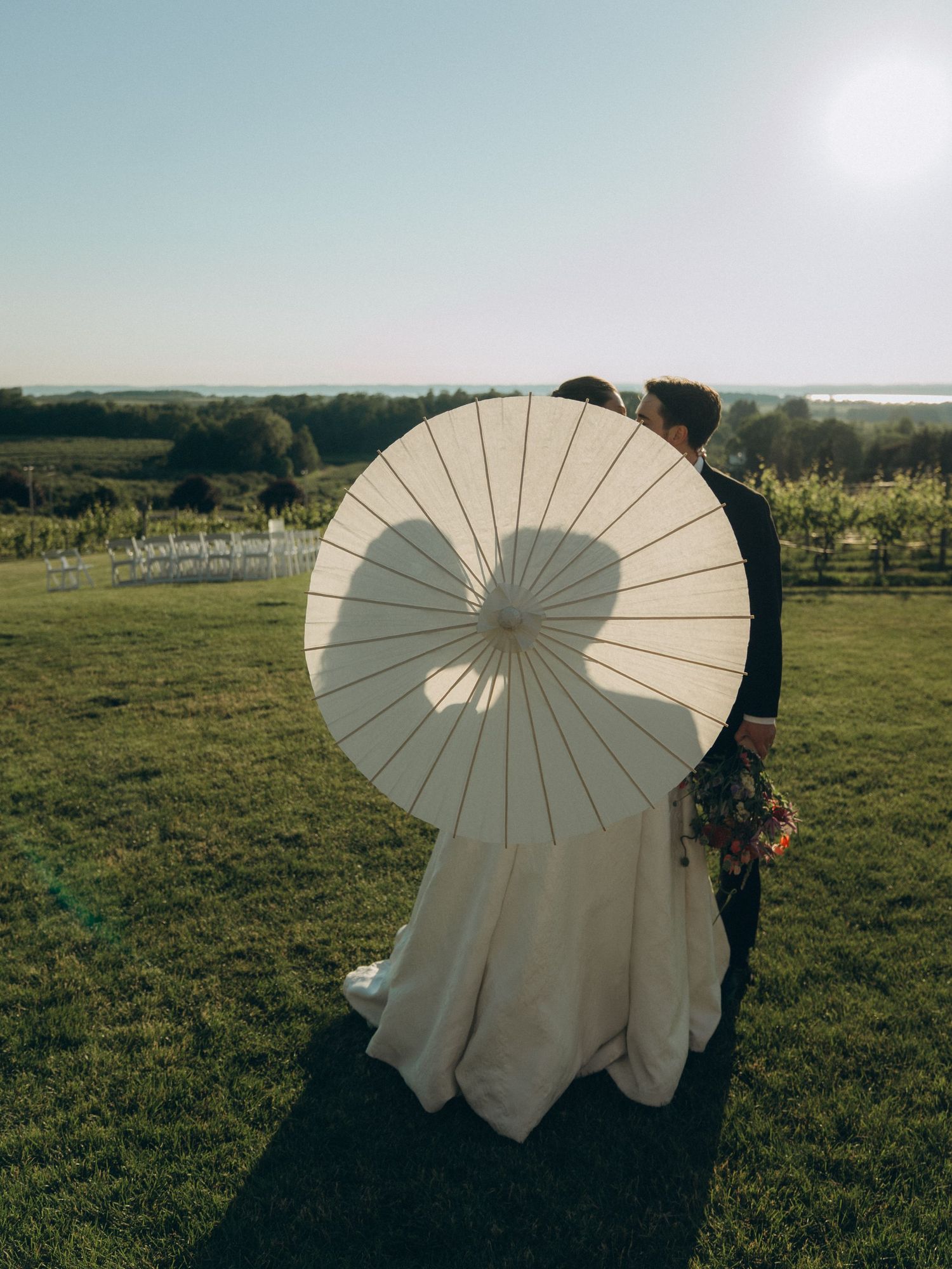 A bride and groom share a romantic kiss behind a white parasol umbrella in a lush vineyard wedding setting.