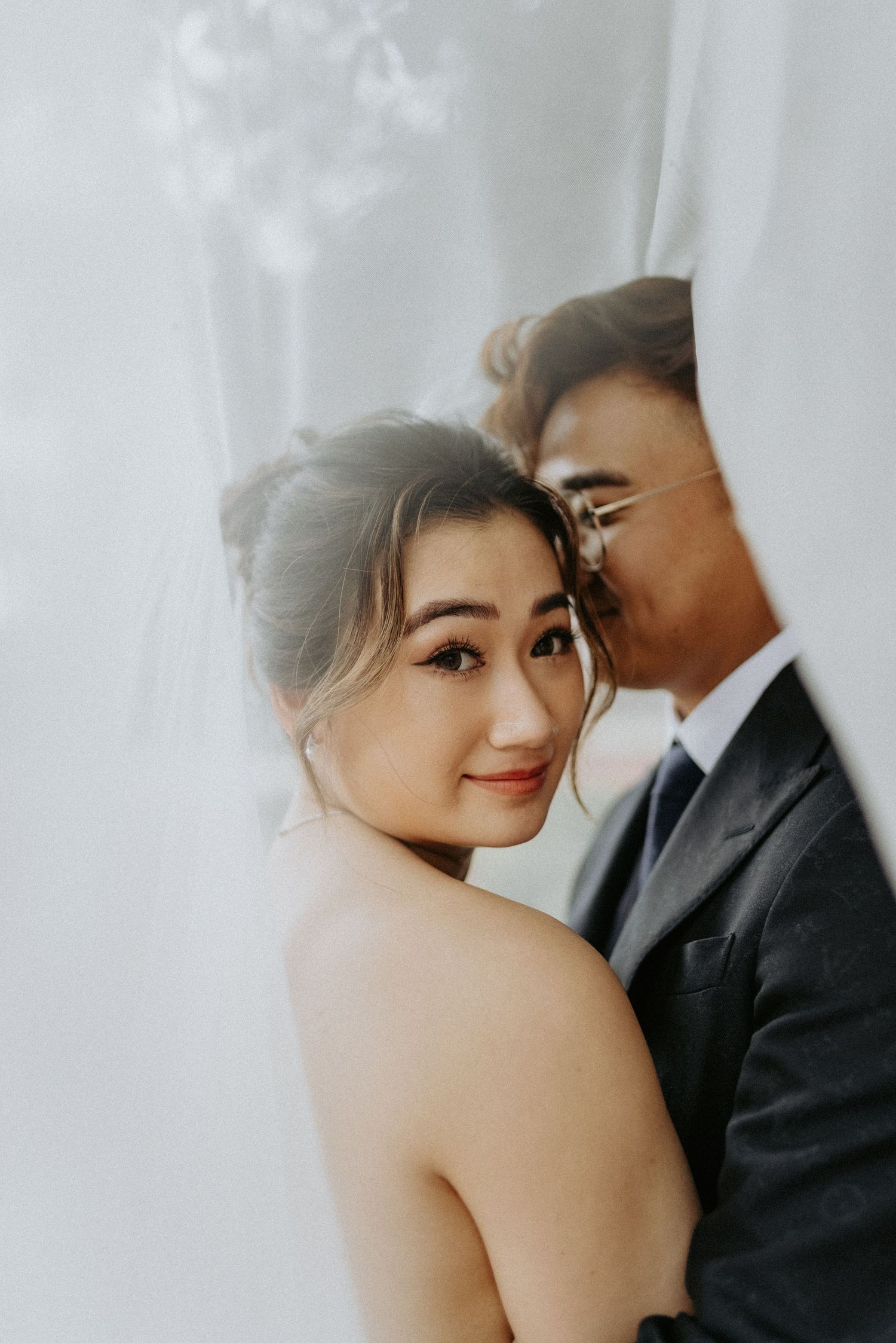 Romantic wedding couple embraces intimately in soft white lighting during professional photoshoot.