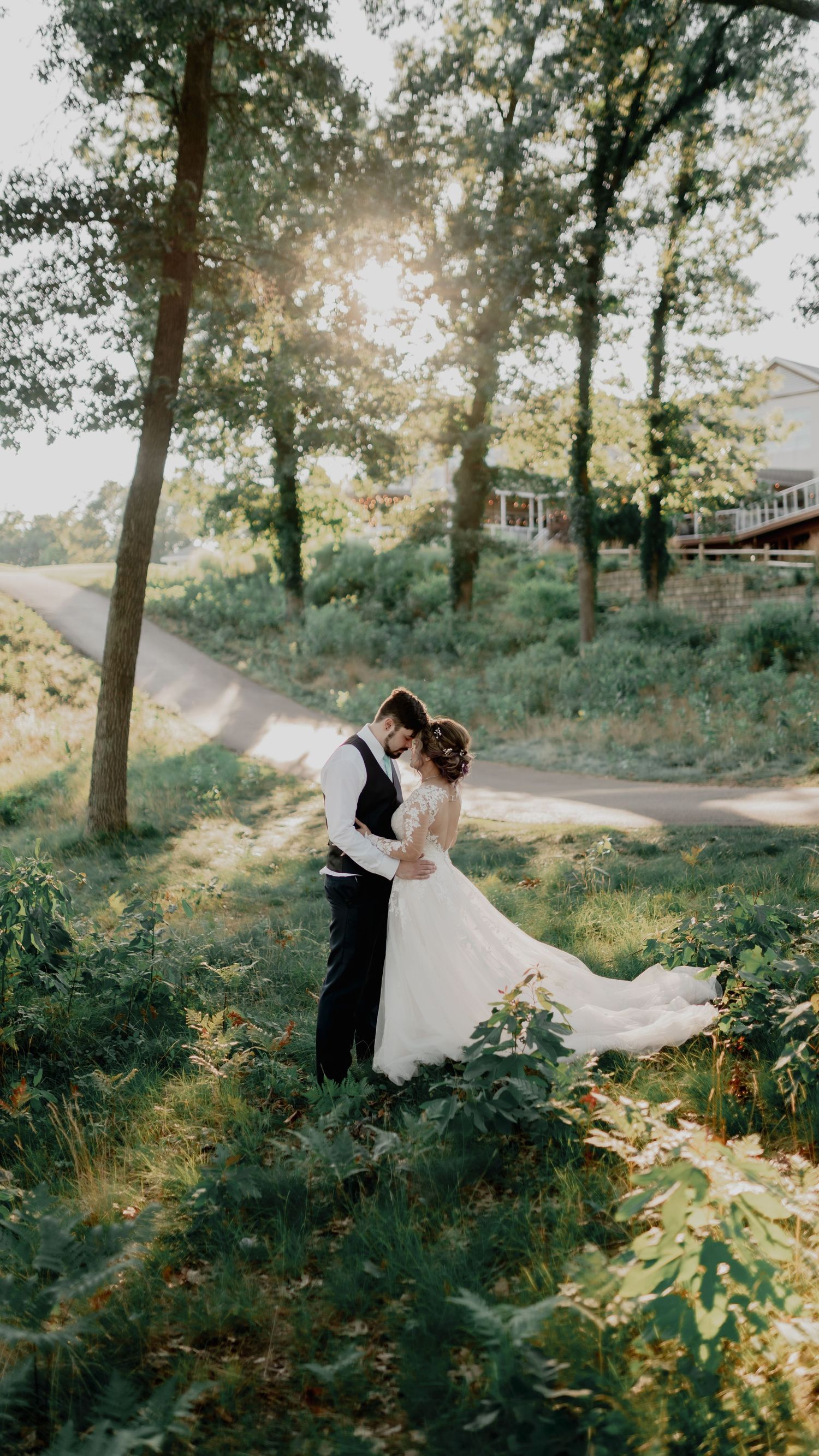 A romantic couple in formal attire embrace amongst sunlit trees and ferns during a woodland photo session.